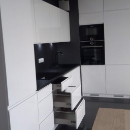 Firma MARCZAK - Nowoczesna, biała kuchnia z czarnym blatem i otwartymi szufladami, widoczne czarne AGD w zabudowie.