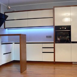 Firma MARCZAK - Nowoczesna zabudowa kuchenna z białymi, lakierowanymi frontami i drewnianymi akcentami w Radomsku, oświetlona listwą LED pod szafkami wiszącymi i lampami sufitowymi.