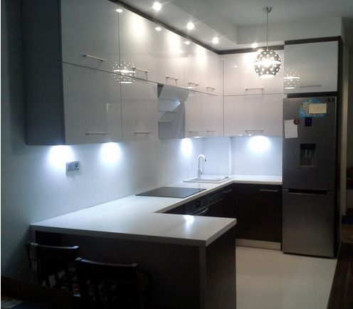 Nowoczesna kuchnia z białymi, błyszczącymi szafkami górnymi i ciemnobrązowymi dolnymi, oświetlona punktowymi lampami podszafkowymi i wiszącą lampą kulą. Widoczny fragment blatu z płytą indukcyjną...