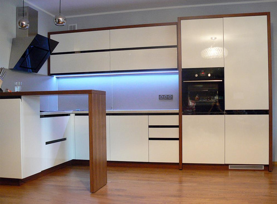 Nowoczesna zabudowa kuchenna z białymi, lakierowanymi frontami i drewnianymi akcentami w Radomsku, oświetlona listwą LED pod szafkami wiszącymi i lampami sufitowymi.