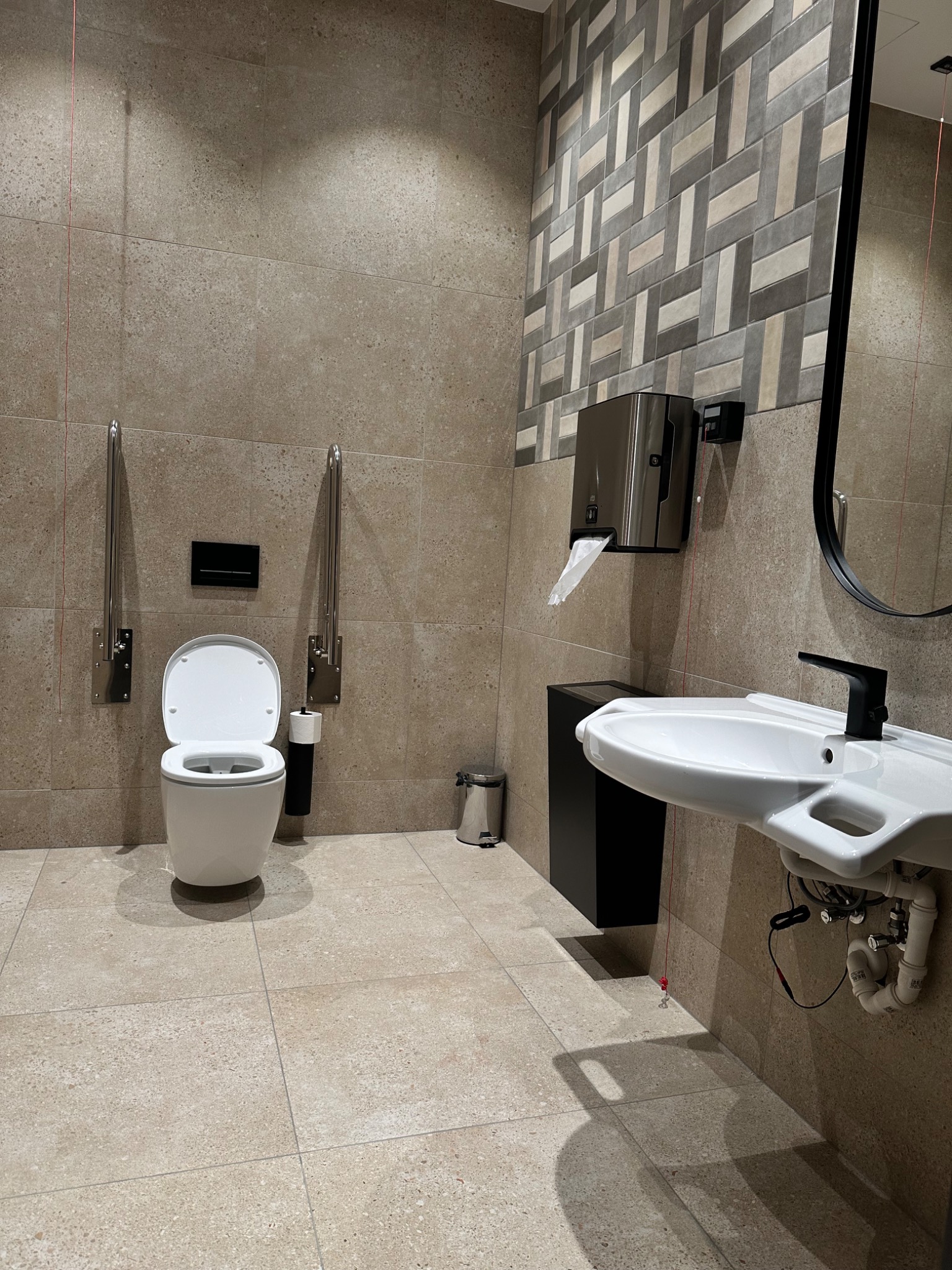 Wykończona łazienka w Katowicach z toaletą dla niepełnosprawnych, umywalką z czarną armaturą i podajnikiem na ręczniki. Beżowe płytki i dekoracyjny pas na ścianie.