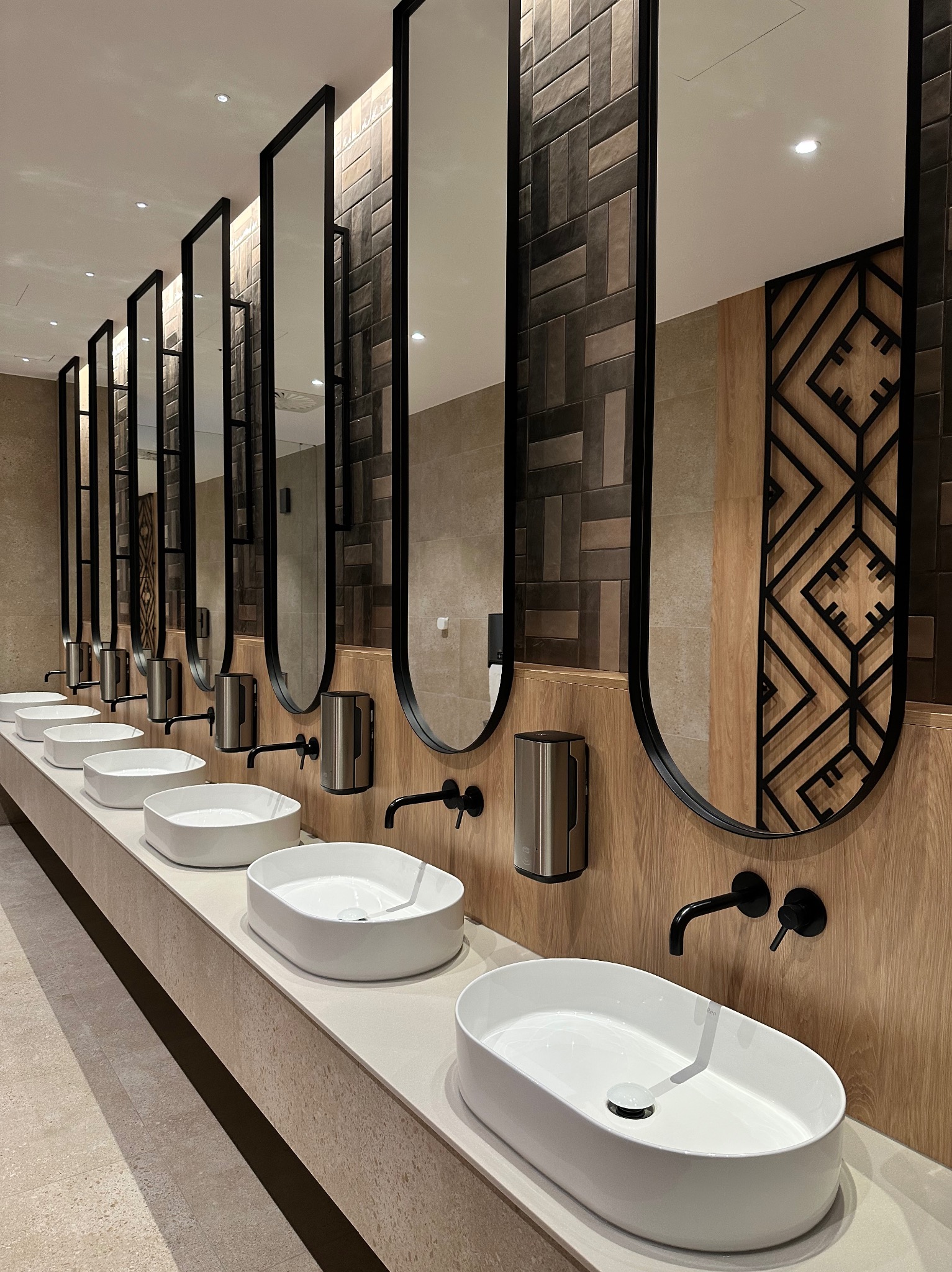 Rząd umywalek w nowoczesnej łazience publicznej z czarnymi bateriami i owalnymi lustrami w czarnych ramach, na tle drewnianej ściany z geometrycznym wzorem.