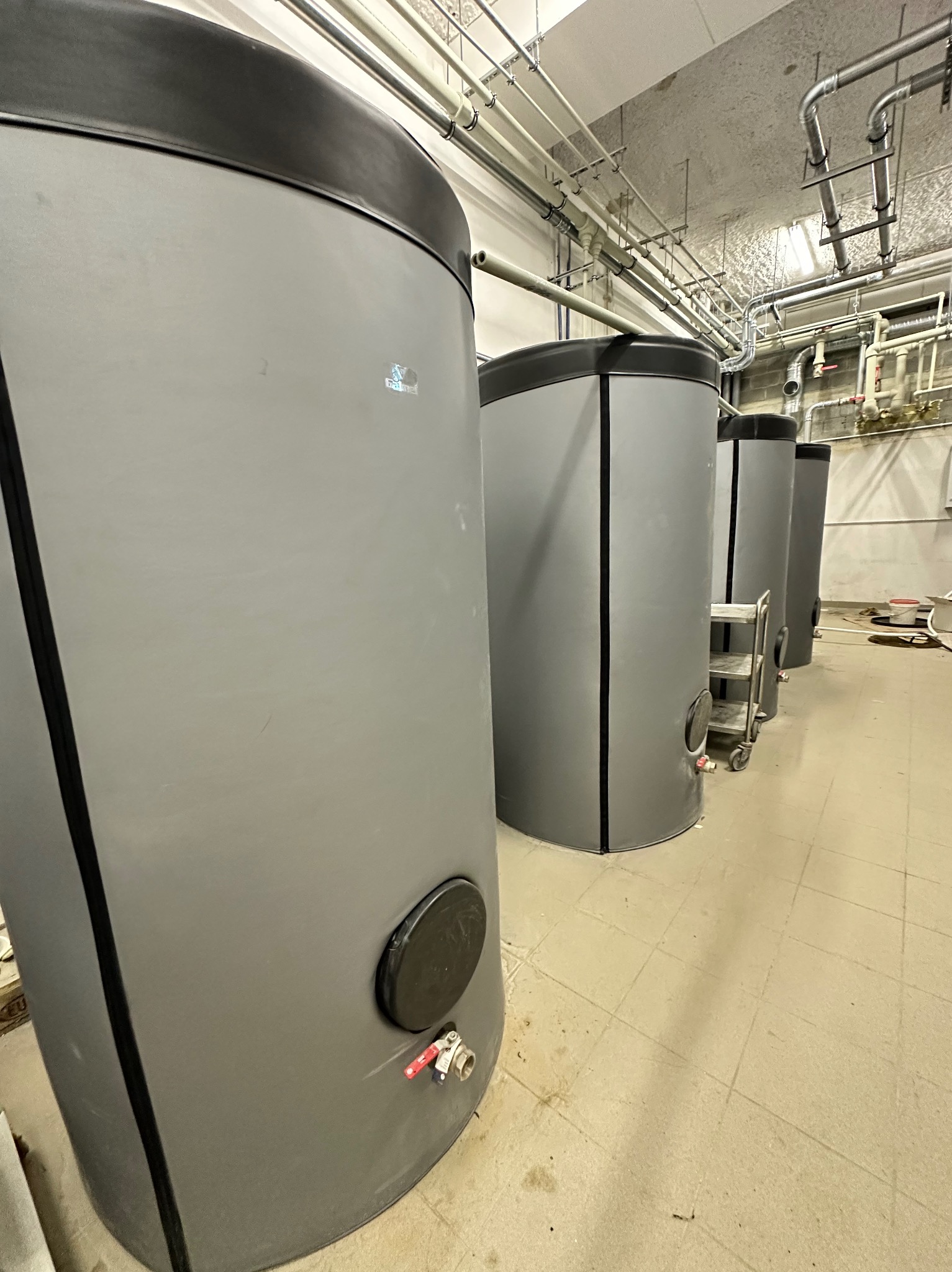 Rząd szarych, pionowych zbiorników z czarnymi pokrywami w pomieszczeniu z rurami pod sufitem. Widoczne zawory i drabinka na kółkach. Ujęcie z dołu.