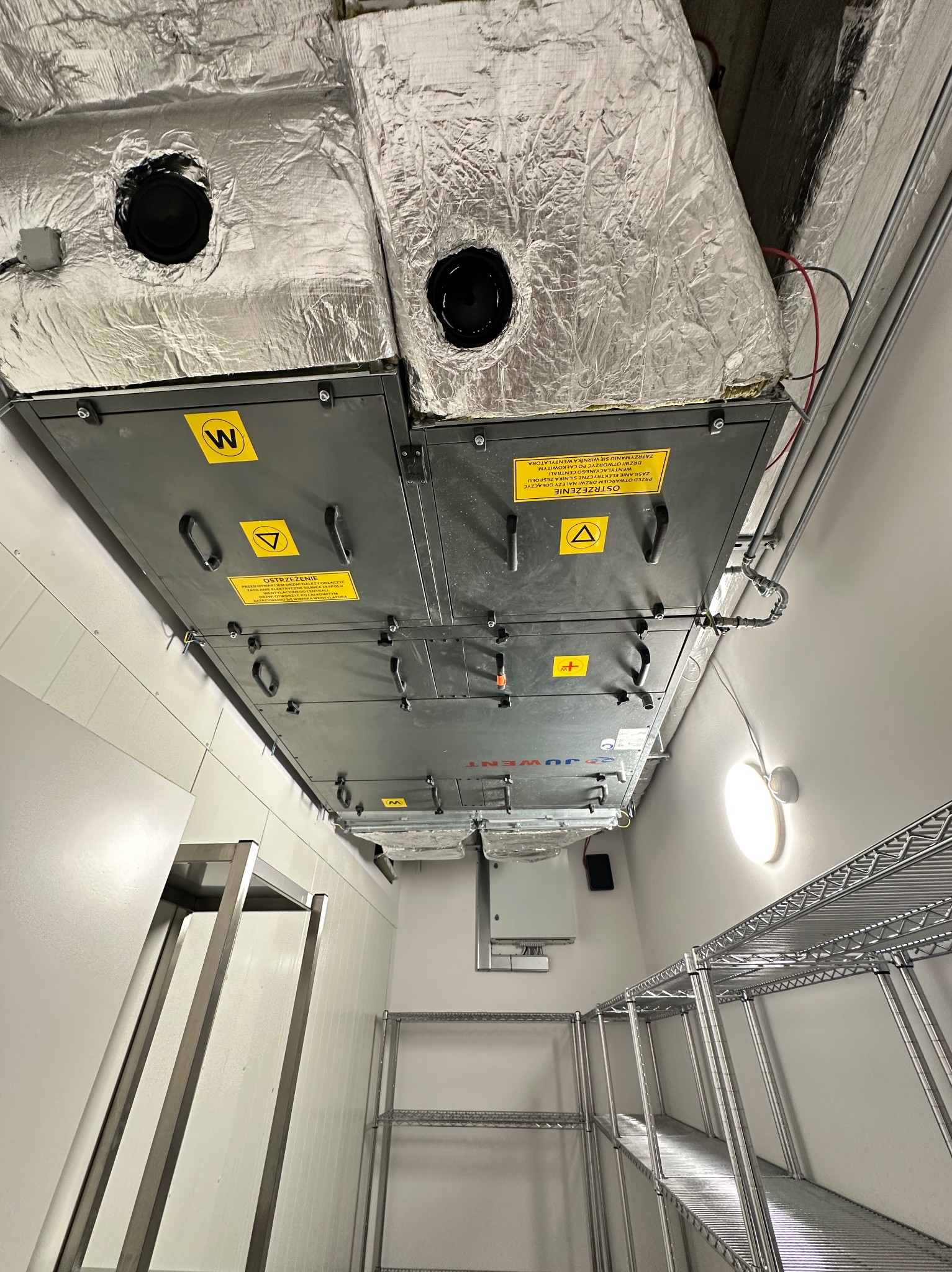 Klimatyzacja przemysłowa Juwent z izolowanymi przewodami wentylacyjnymi, widoczna w pomieszczeniu z metalowymi regałami i oświetleniem sufitowym.