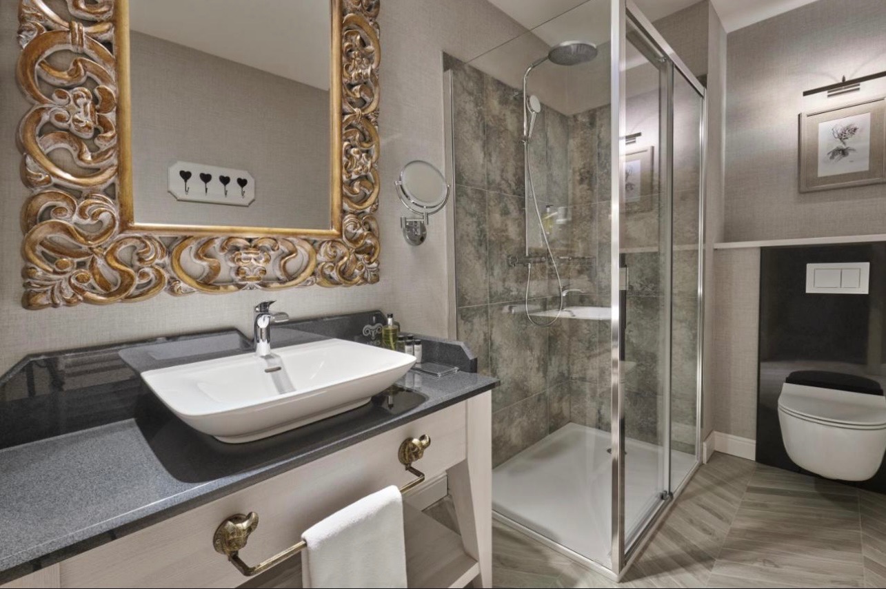 Elegancka łazienka z kabiną prysznicową, umywalką nablatową i ozdobnym lustrem w złotej ramie. Widoczne detale wykończenia i armatura.
