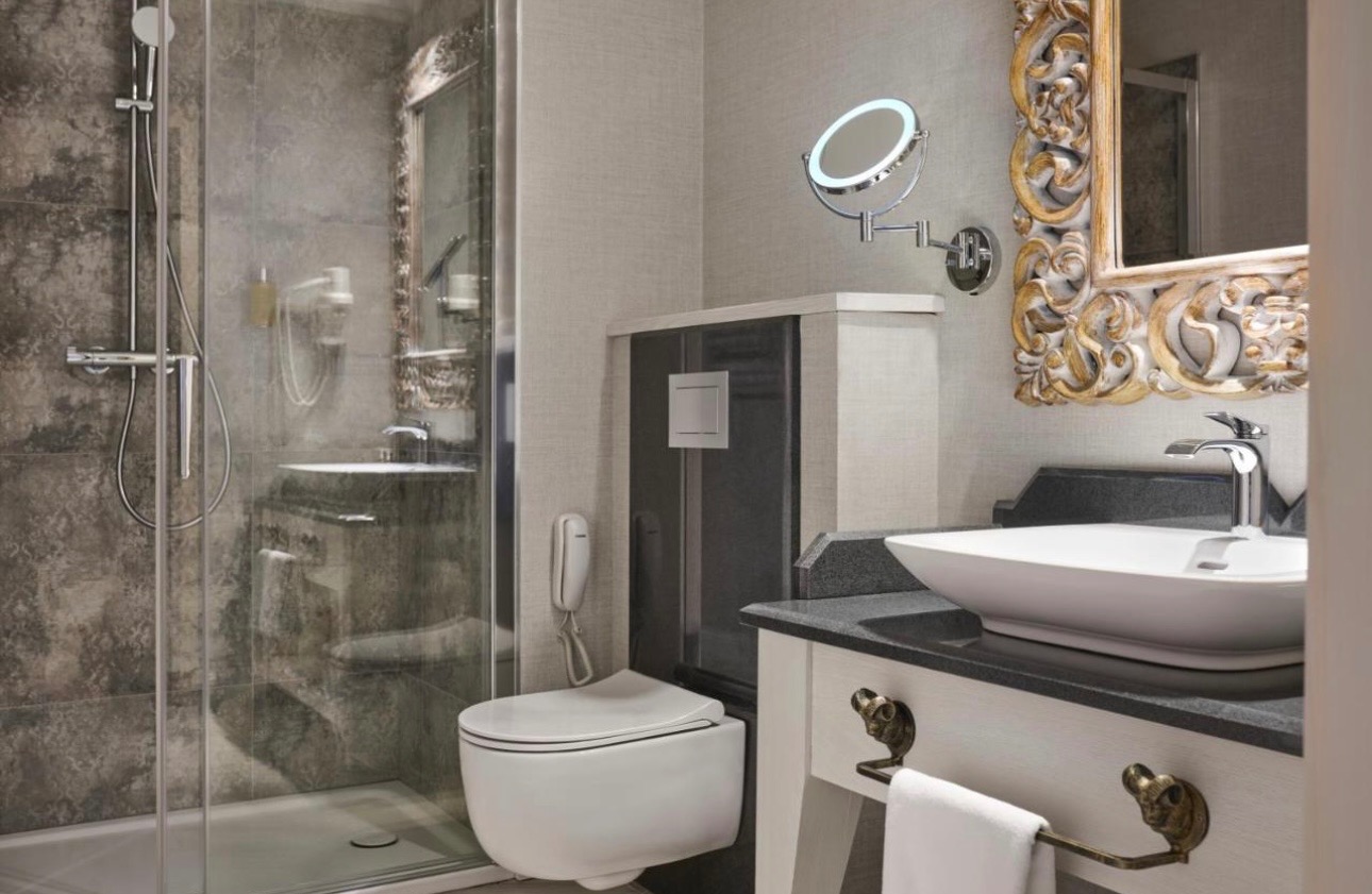 Elegancka łazienka z kabiną prysznicową, umywalką na blacie z ozdobnymi uchwytami i toaletą podwieszaną. Lustro z podświetleniem i dekoracyjna rama lustra.