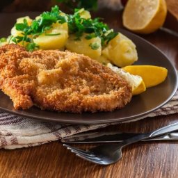 PRO QUALITY GROUP Sp. z o.o. - Sznycel w złocistej panierce z ziemniakami posypanymi natką pietruszki, szparagami i cytryną na brązowym talerzu, obok sztućce i miseczka z sałatką.