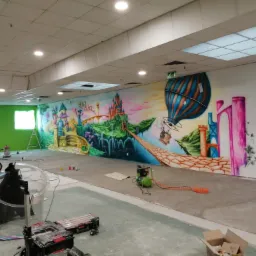 Wnętrze biura z pomalowaną ścianą w fantazyjny motyw zamek, balon i elementy przyrody, widoczne narzędzia malarskie i kompresor, zielone ściany w tle.