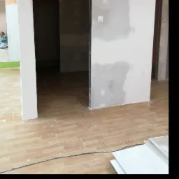 Wnętrze w trakcie prac wykończeniowych: ściany z płyt kartonowo-gipsowych z widocznymi łączeniami, podłoga wyłożona panelami imitującymi drewno, widoczny kabel elektryczny i stos płyt gipsowych...