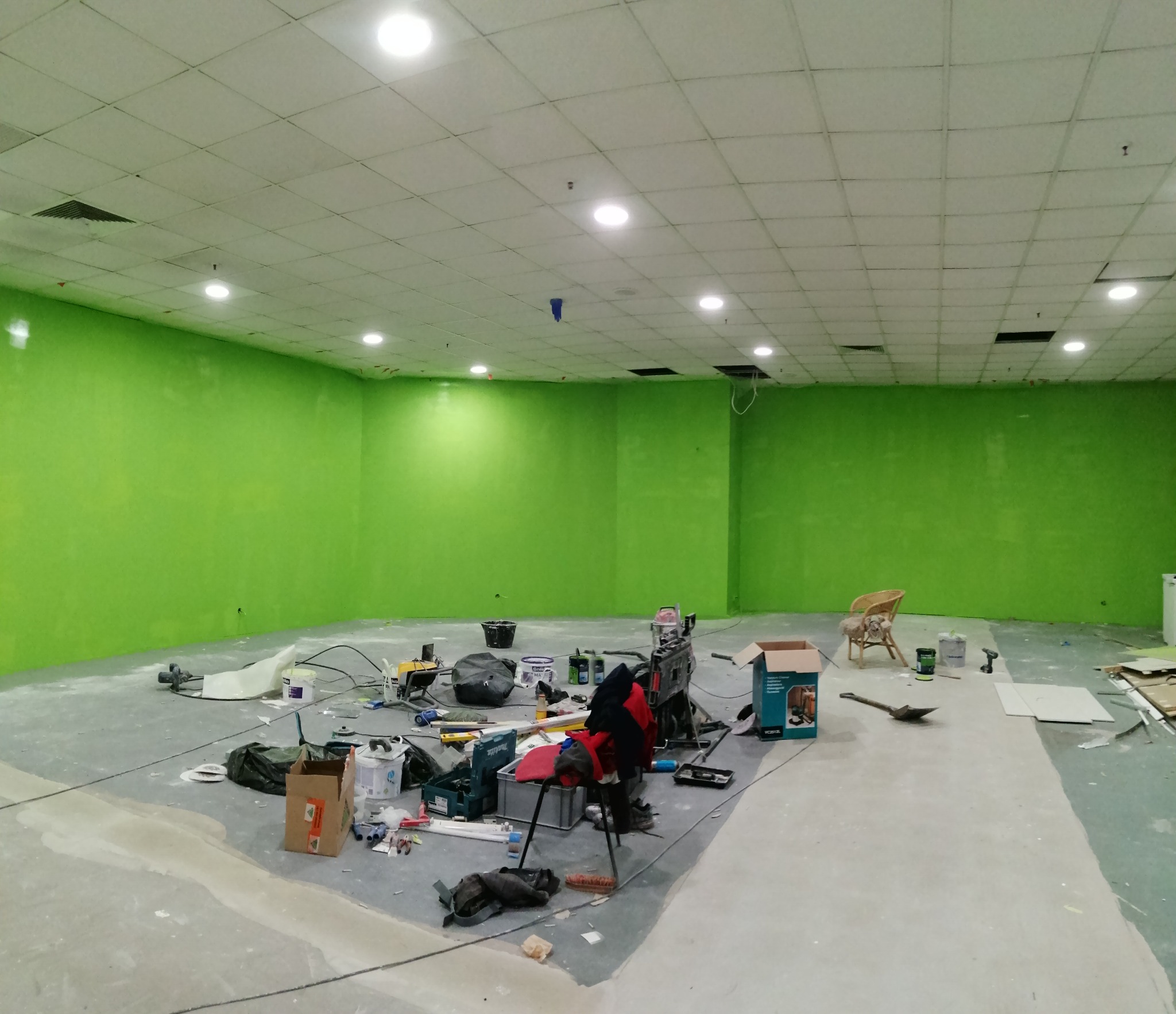 Wnętrze biura w trakcie malowania na jaskrawy zielony kolor, widoczne narzędzia malarskie, wiadro z farbą, wałki, pędzle, drabina oraz krzesło wiklinowe w rogu pomieszczenia.