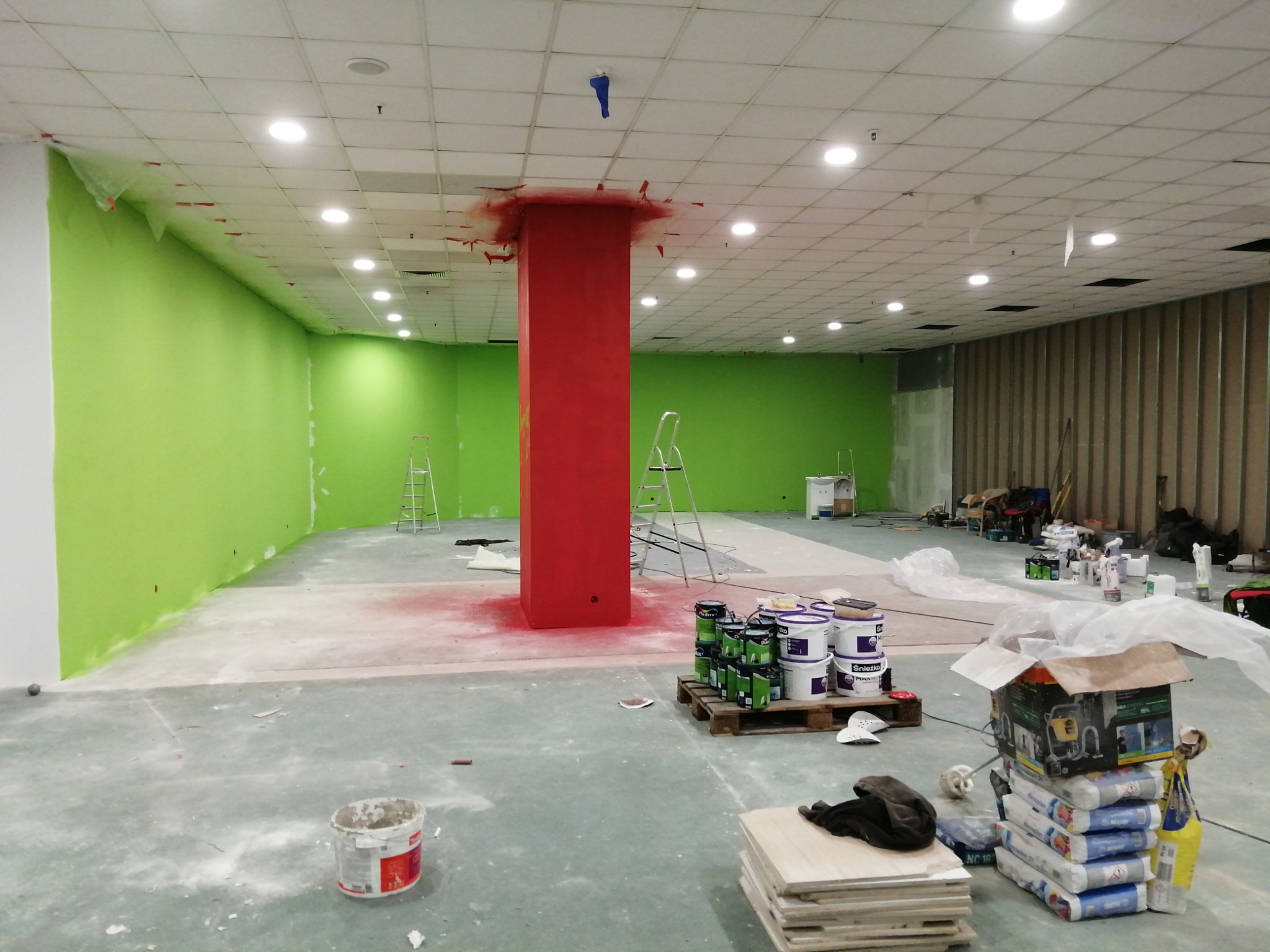 Wnętrze biura w trakcie malowania: ściany pomalowane na jasnozielono, kolumna w intensywnym czerwonym kolorze, zabezpieczona folią, widoczne puszki z farbą, drabina i narzędzia malarskie.