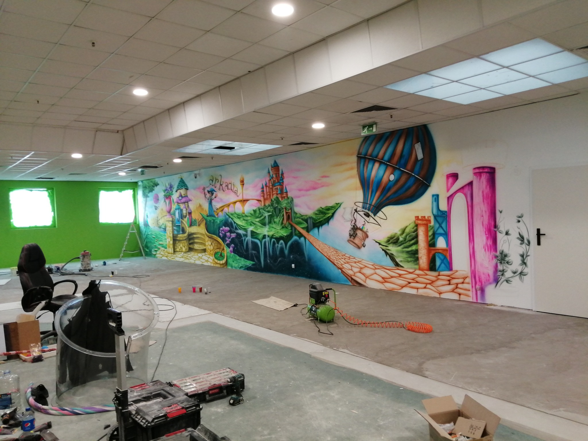 Wnętrze biura z pomalowaną ścianą w fantazyjny motyw zamek, balon i elementy przyrody, widoczne narzędzia malarskie i kompresor, zielone ściany w tle.