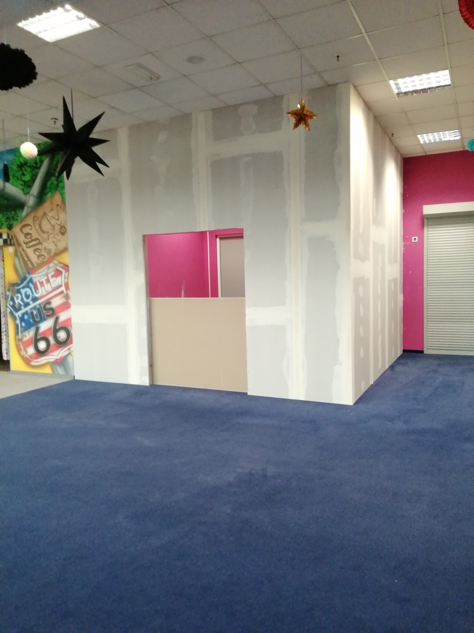 Wnętrze w trakcie prac wykończeniowych: montaż płyt kartonowo-gipsowych na ścianach w pomieszczeniu z niebieską wykładziną, widoczny fragment ściany pomalowanej na różowo i dekoracje sufitowe...