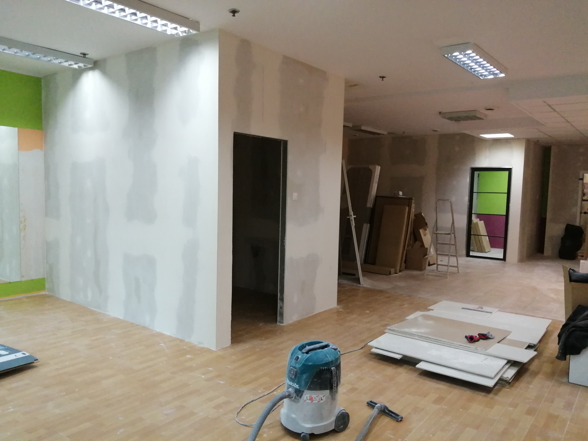 Wnętrze biura w trakcie prac wykończeniowych: szpachlowane ściany, przygotowane płyty gipsowo-kartonowe, odkurzacz przemysłowy Makita i narzędzia na parkiecie. Widoczne otwarte przejście...