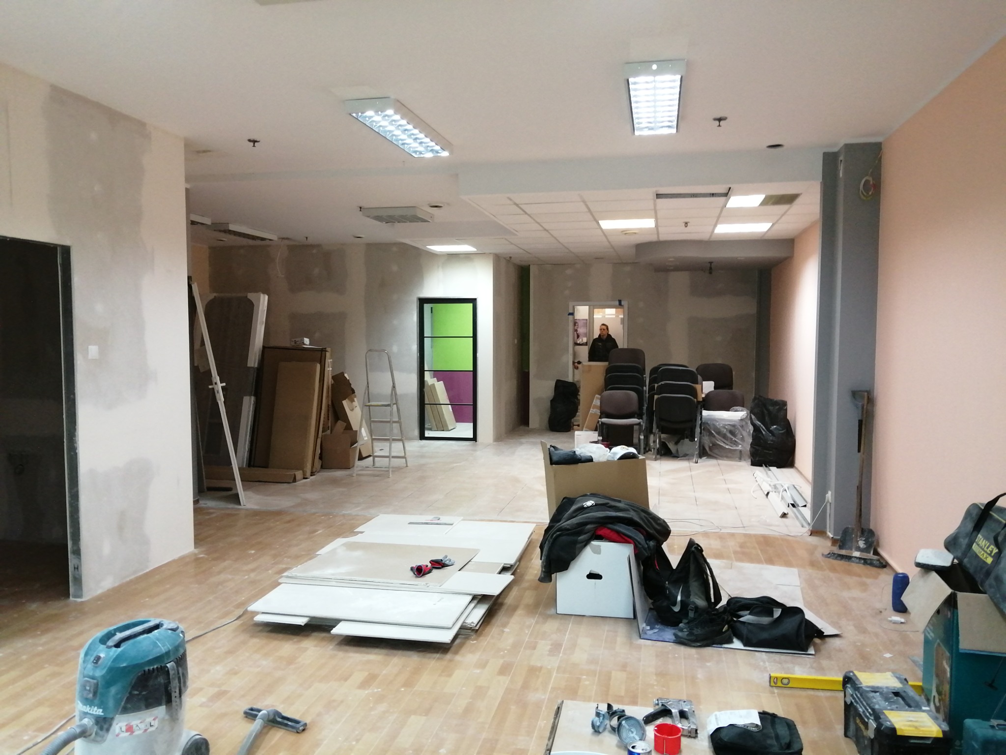 Wnętrze biura w trakcie prac wykończeniowych; widoczne płyty karton gips, narzędzia, odkurzacz przemysłowy oraz przygotowane pomieszczenie z krzesłami.