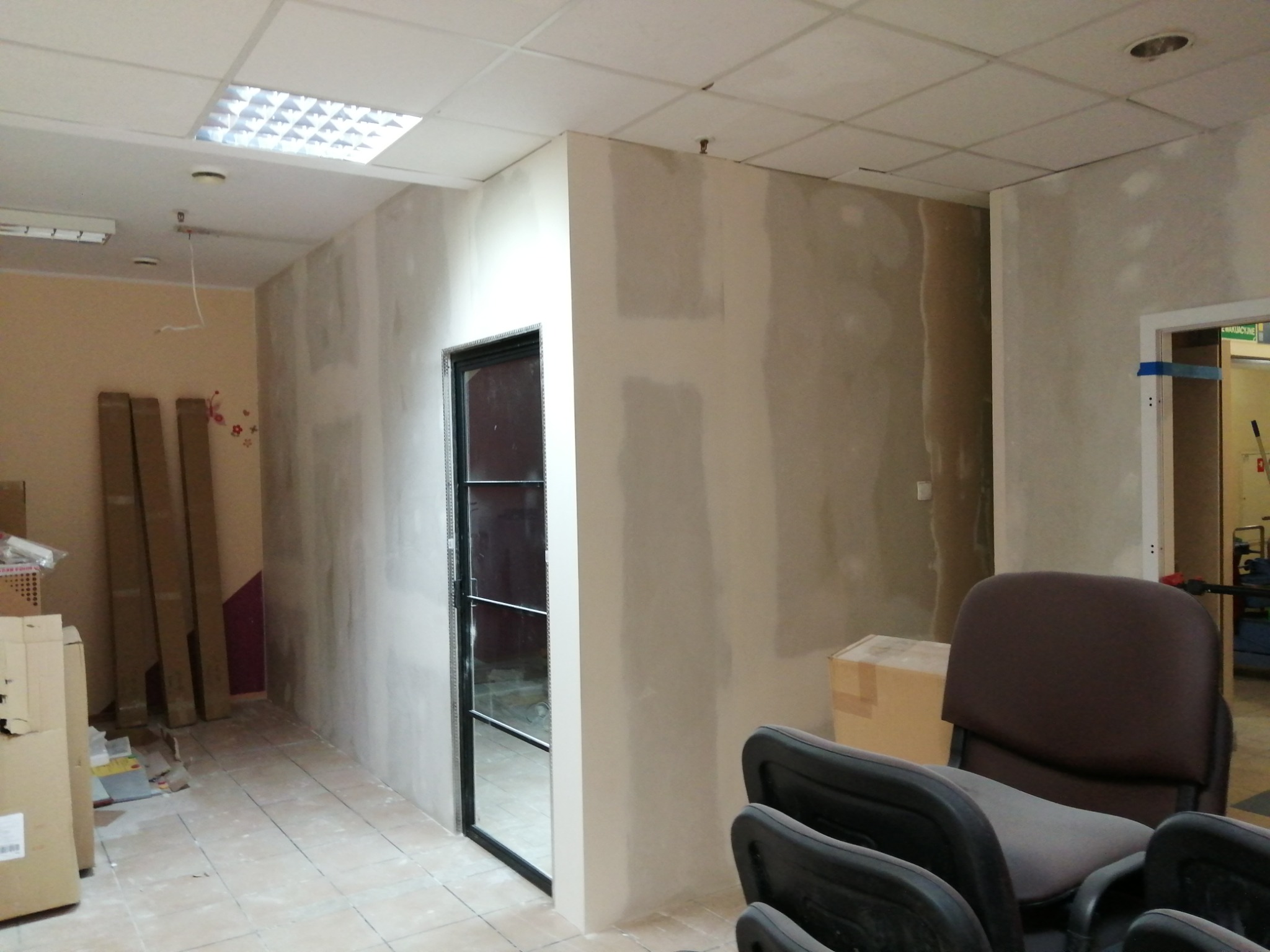 Biuro w trakcie prac wykończeniowych: ściany po szpachlowaniu, przygotowane do malowania, widoczna nowa ościeżnica drzwiowa, kartony i krzesła biurowe w pomieszczeniu.