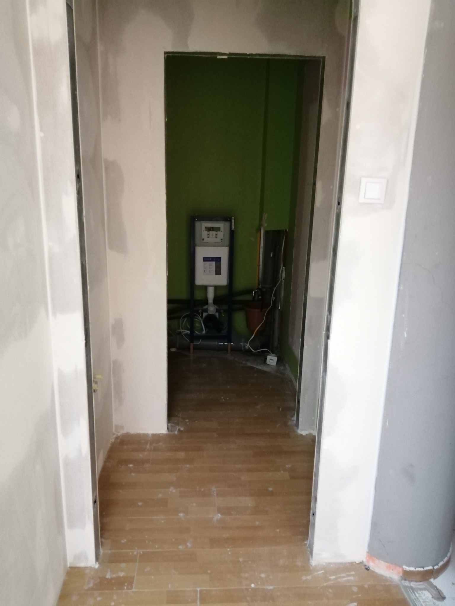 Wnętrze w trakcie prac wykończeniowych: widok przez otwór drzwiowy na zieloną ścianę z zamontowanym wodomierzem, surowe ściany i podłoga z panelami w trakcie układania.