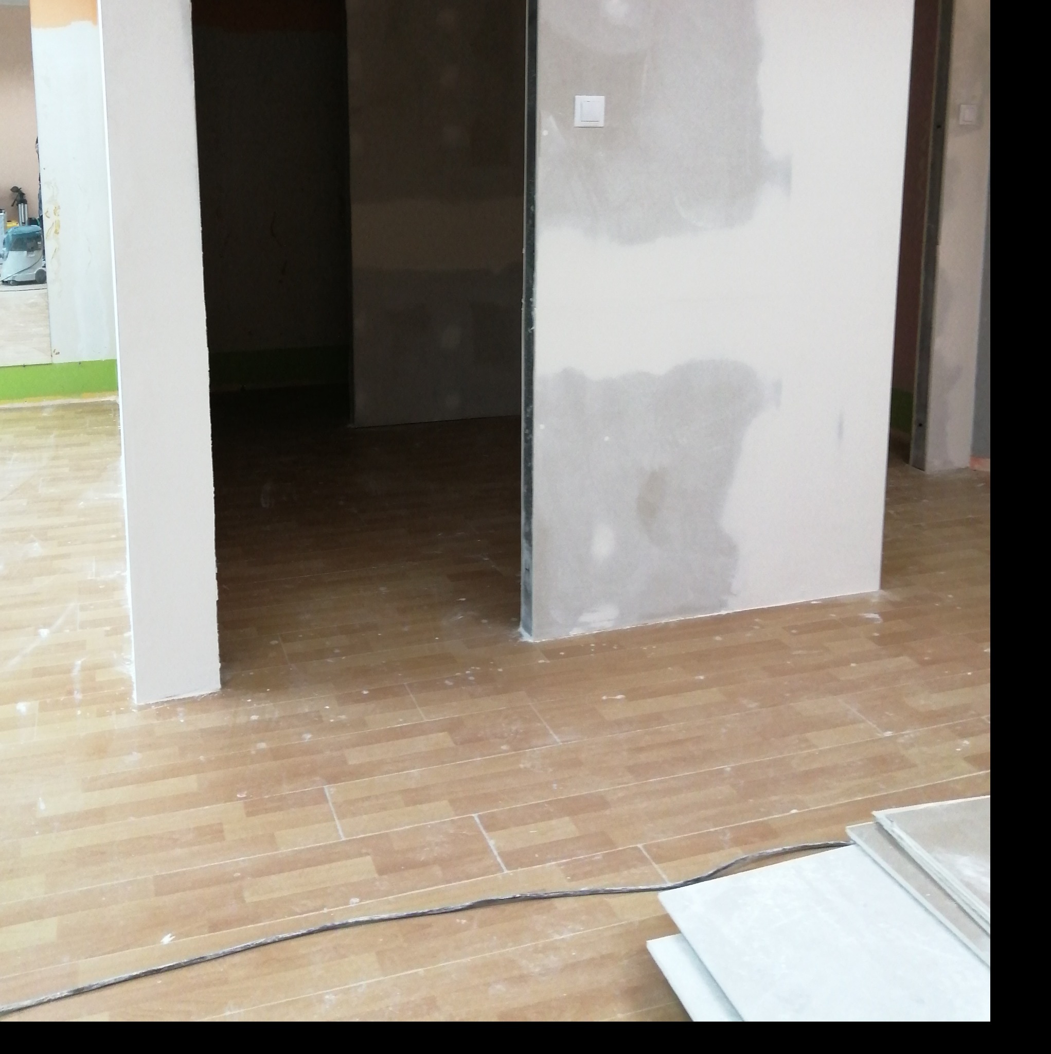 Wnętrze w trakcie prac wykończeniowych: ściany z płyt kartonowo-gipsowych z widocznymi łączeniami, podłoga wyłożona panelami imitującymi drewno, widoczny kabel elektryczny i stos płyt gipsowych...