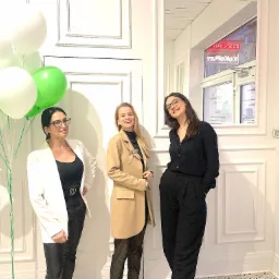 Trzy kobiety ubrane elegancko stoją na tle białej ściany z dekoracyjnymi panelami, obok okna z neonem 'KOKOLS' i napisem 'NIERUCHOMOŚCI'; po lewej stronie bukiet zielonych i białych balonów.