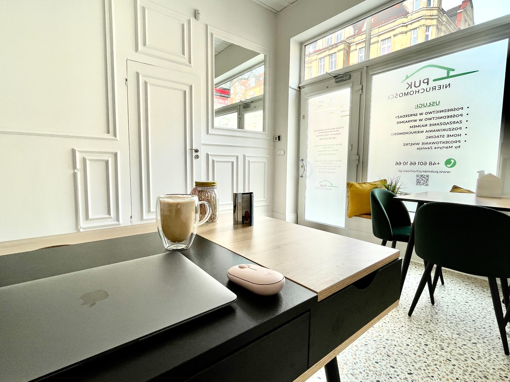 Biuro nieruchomości z laptopem, myszką i kawą na biurku; w tle okno z logo firmy i widok na budynek.