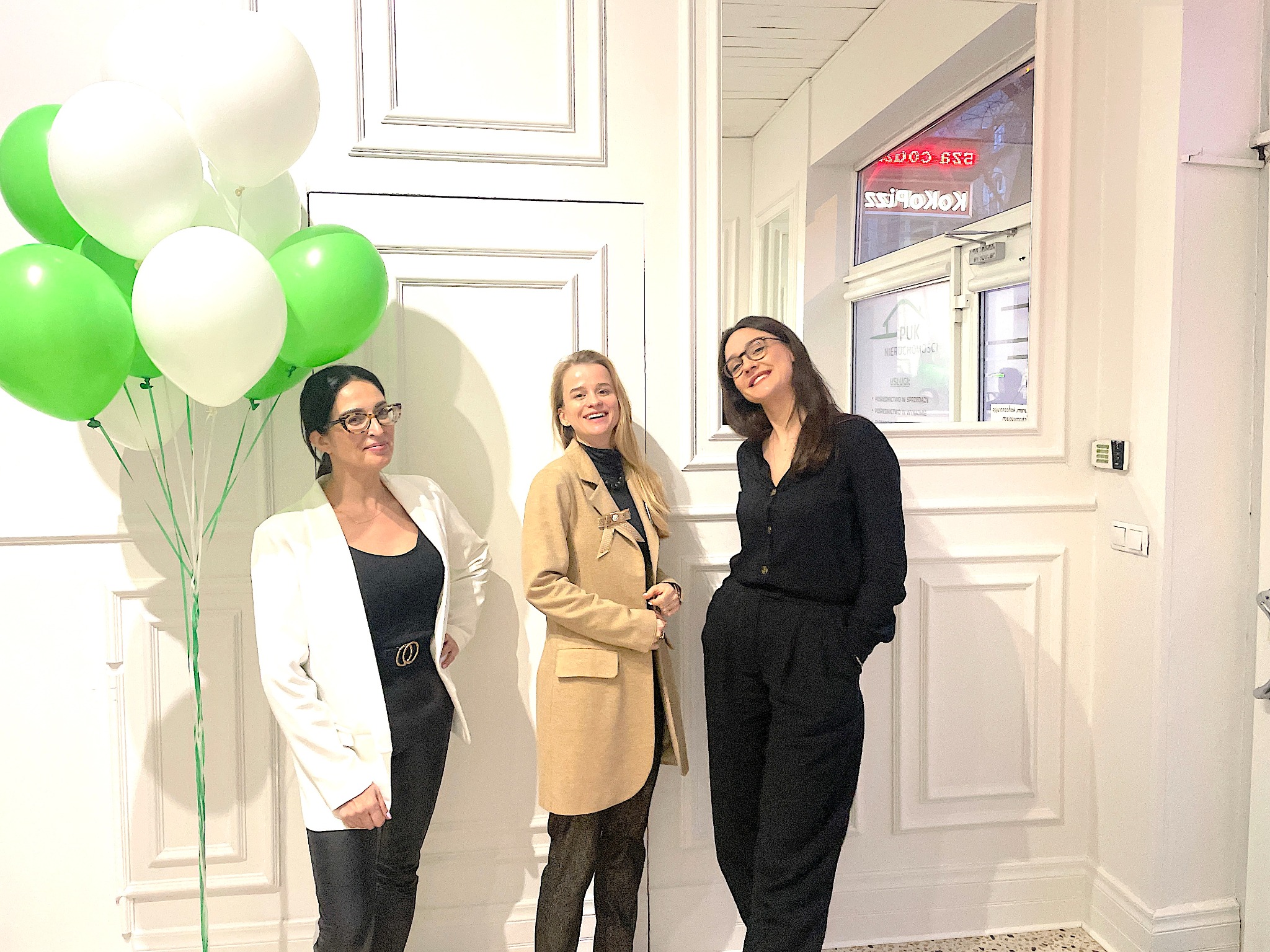 Trzy kobiety ubrane elegancko stoją na tle białej ściany z dekoracyjnymi panelami, obok okna z neonem 'KOKOLS' i napisem 'NIERUCHOMOŚCI'; po lewej stronie bukiet zielonych i białych balonów.