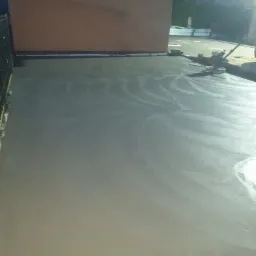 Świeżo wylana, gładka powierzchnia betonowa na zewnątrz budynku, widoczne ślady obróbki mechaniczną pacą, w tle fragment budynku i ogrodzenia.