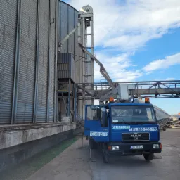 Niebieski samochód ciężarowy z wysięgnikiem zaparkowany obok metalowych silosów zbożowych, widoczny numer telefonu i logo firmy Wiesecker, podpora stabilizująca pojazd.