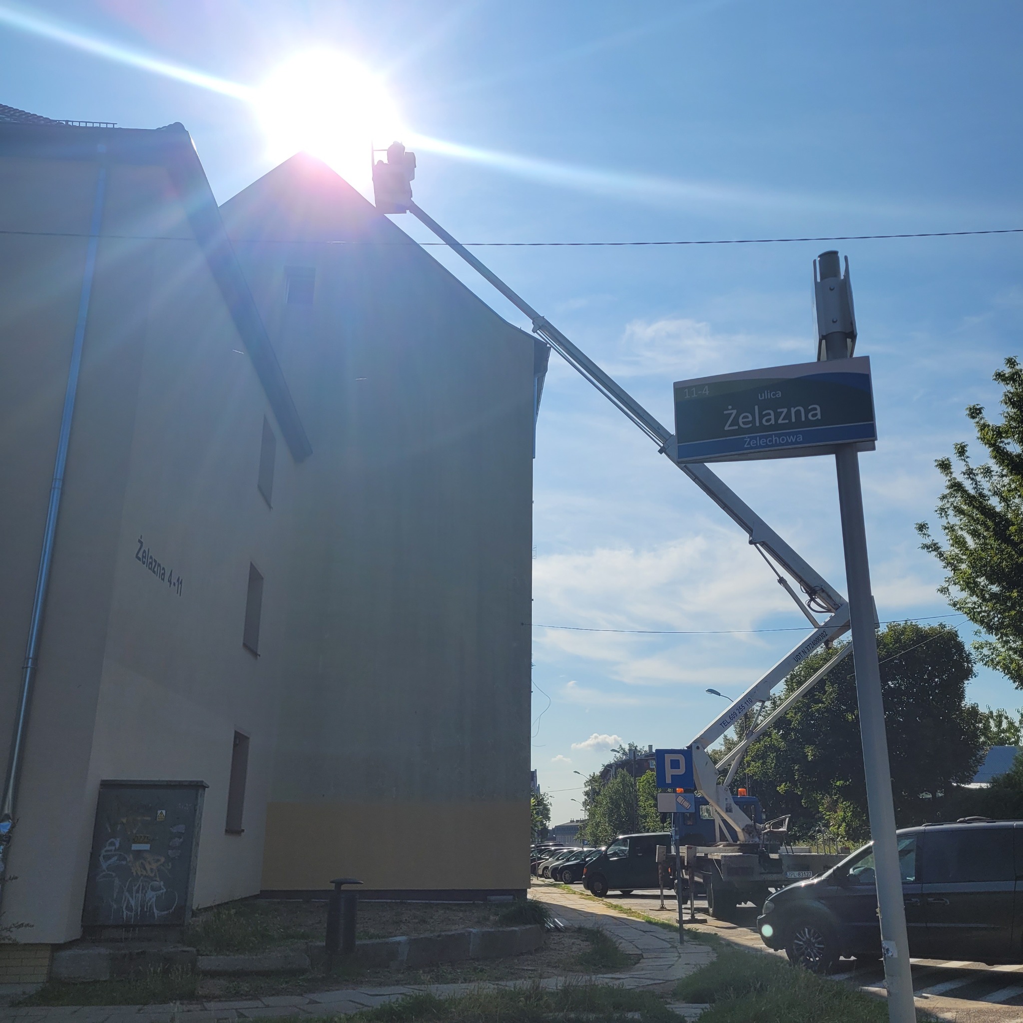 Dwa podnośniki koszowe pracują na wysokości budynku przy ulicy Żelaznej w słoneczny dzień, jeden z nich podnosi pracownika na dach, drugi stoi na ulicy przy znaku drogowym.