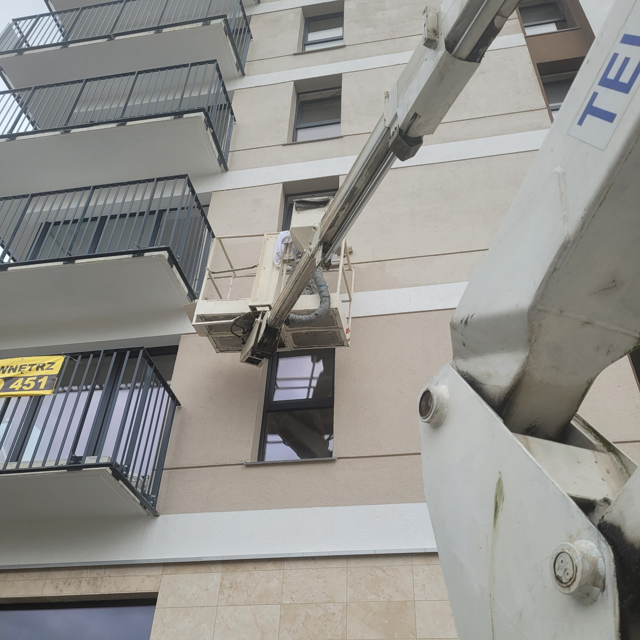 Podnośnik koszowy przy elewacji budynku wielorodzinnego podczas prac montażowych lub konserwacyjnych okien.