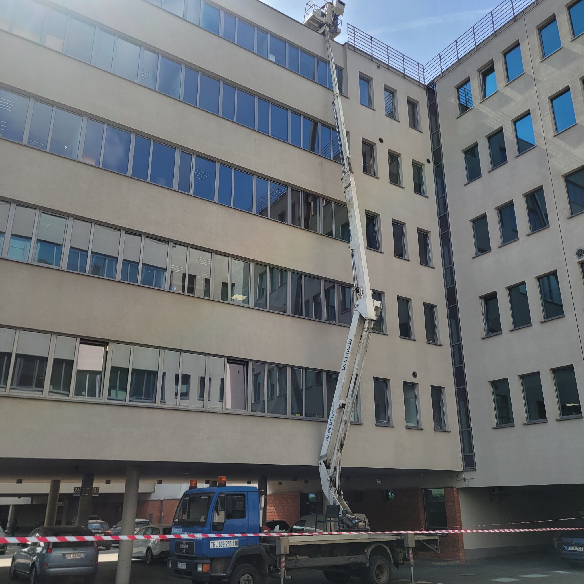 Praca na wysokości przy budynku biurowym z użyciem wysięgnika zamontowanego na samochodzie ciężarowym, widoczne okna budynku i błękitne niebo.