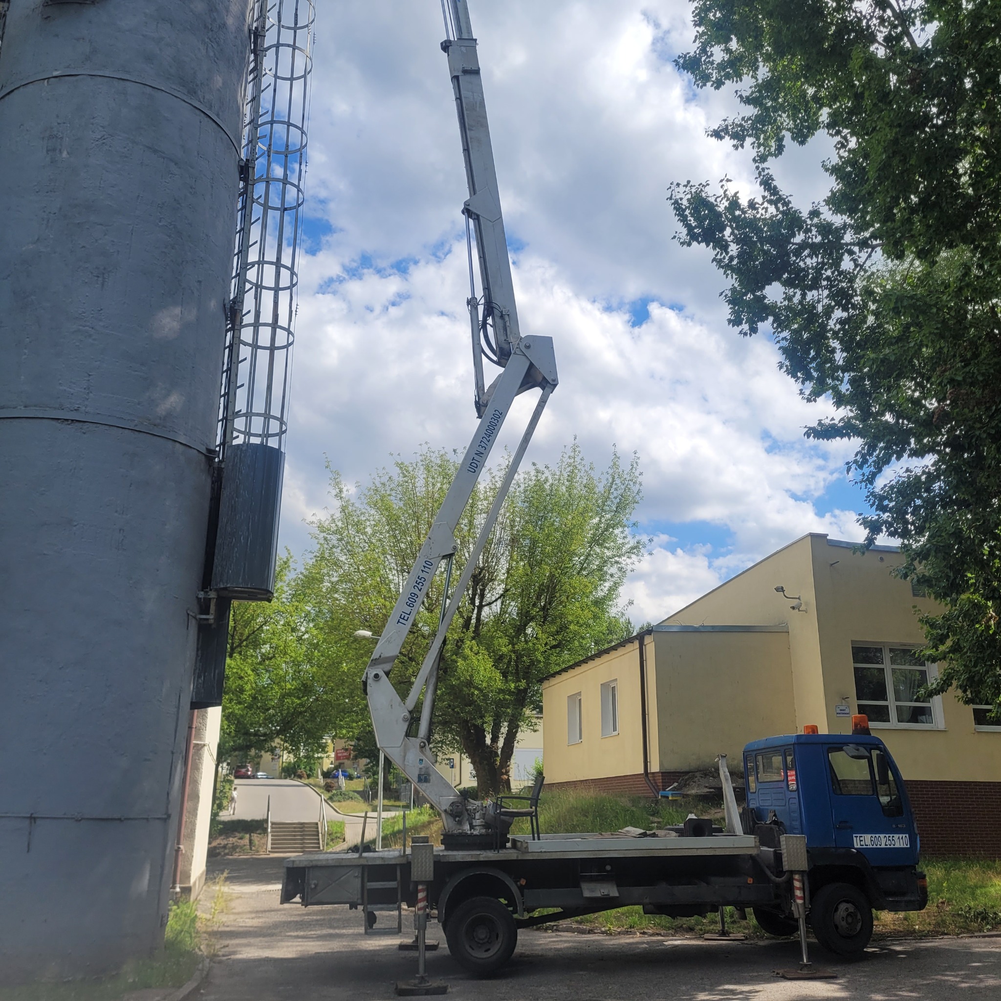 Niebieski samochód z wysięgnikiem teleskopowym ustawiony obok szarego, wysokiego silosu z drabinką, na tle błękitnego nieba z chmurami i budynku administracyjnego.