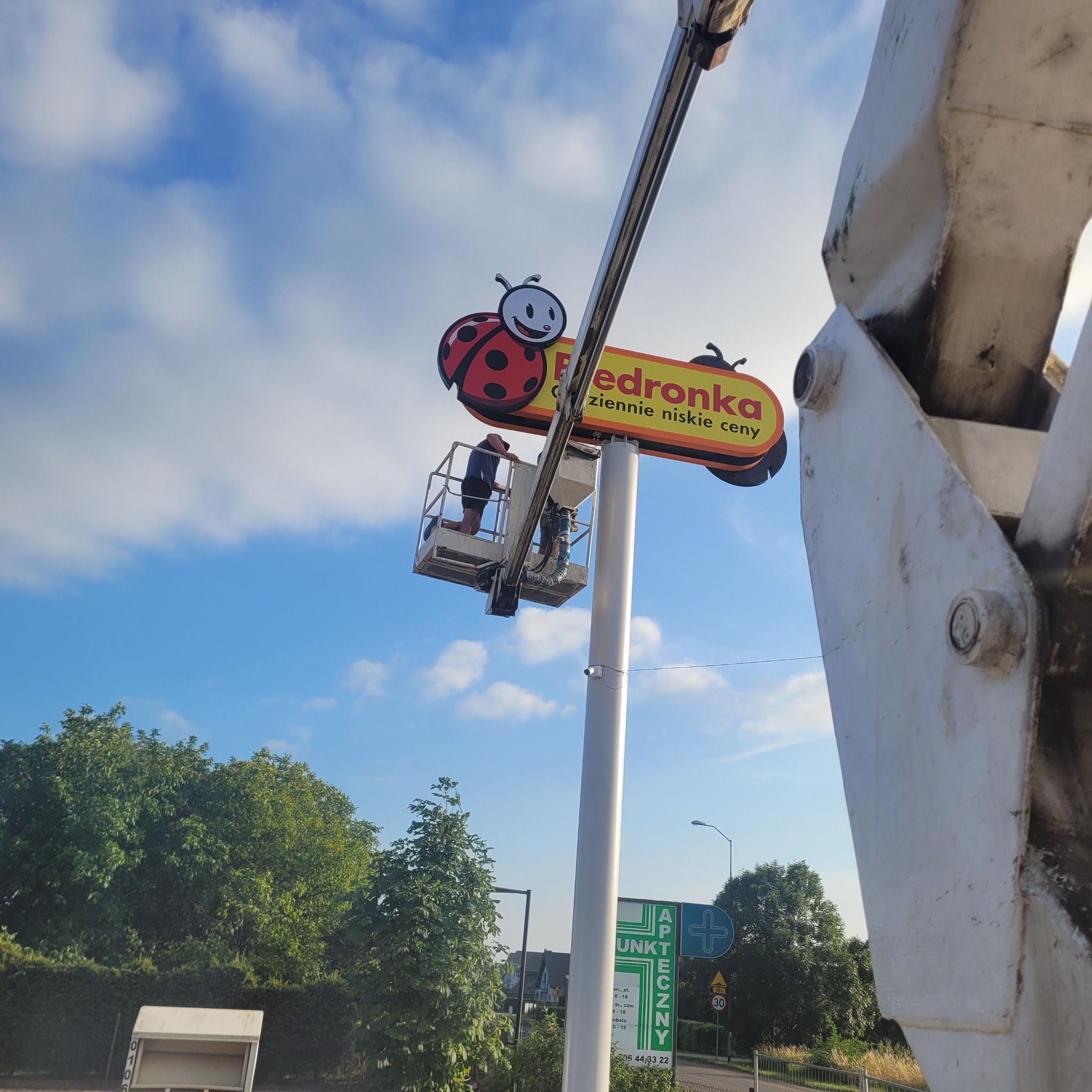 Montaż lub serwis szyldu reklamowego sklepu z użyciem podnośnika koszowego, dwóch pracowników w koszu na wysokości, błękitne niebo w tle.