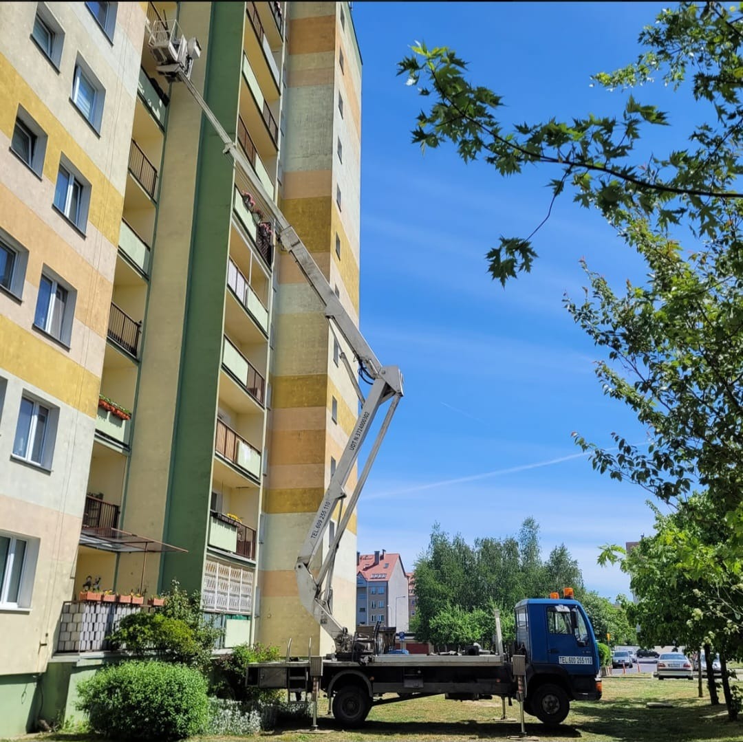 Praca na wysokościach: Dwóch pracowników w koszu podnośnika teleskopowego na tle żółto-zielonego bloku mieszkalnego, błękitne niebo w tle, widoczne fragmenty drzew.
