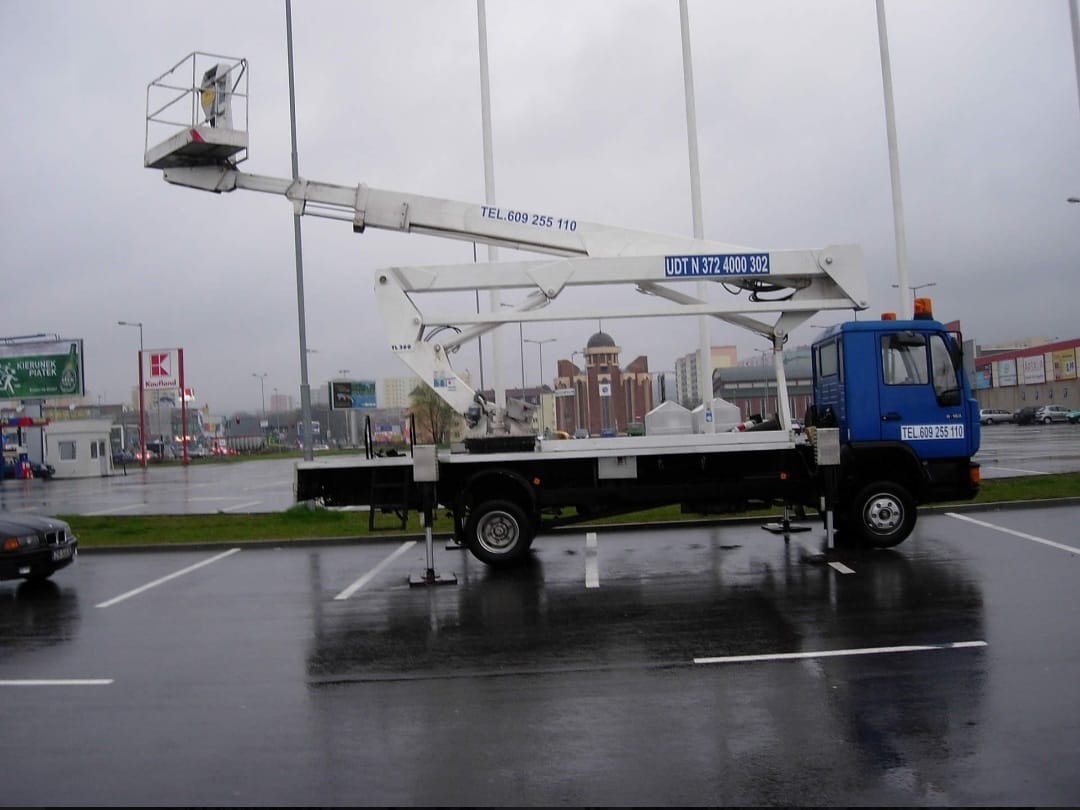 Niebieska ciężarówka z wysięgnikiem na parkingu w Szczecinie w pochmurny dzień. Na wysięgniku widoczny kosz. Na parkingu mokro po deszczu.