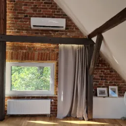 Klimatyzator ścienny zamontowany na ceglanej ścianie poddasza z drewnianymi belkami stropowymi i oknami.