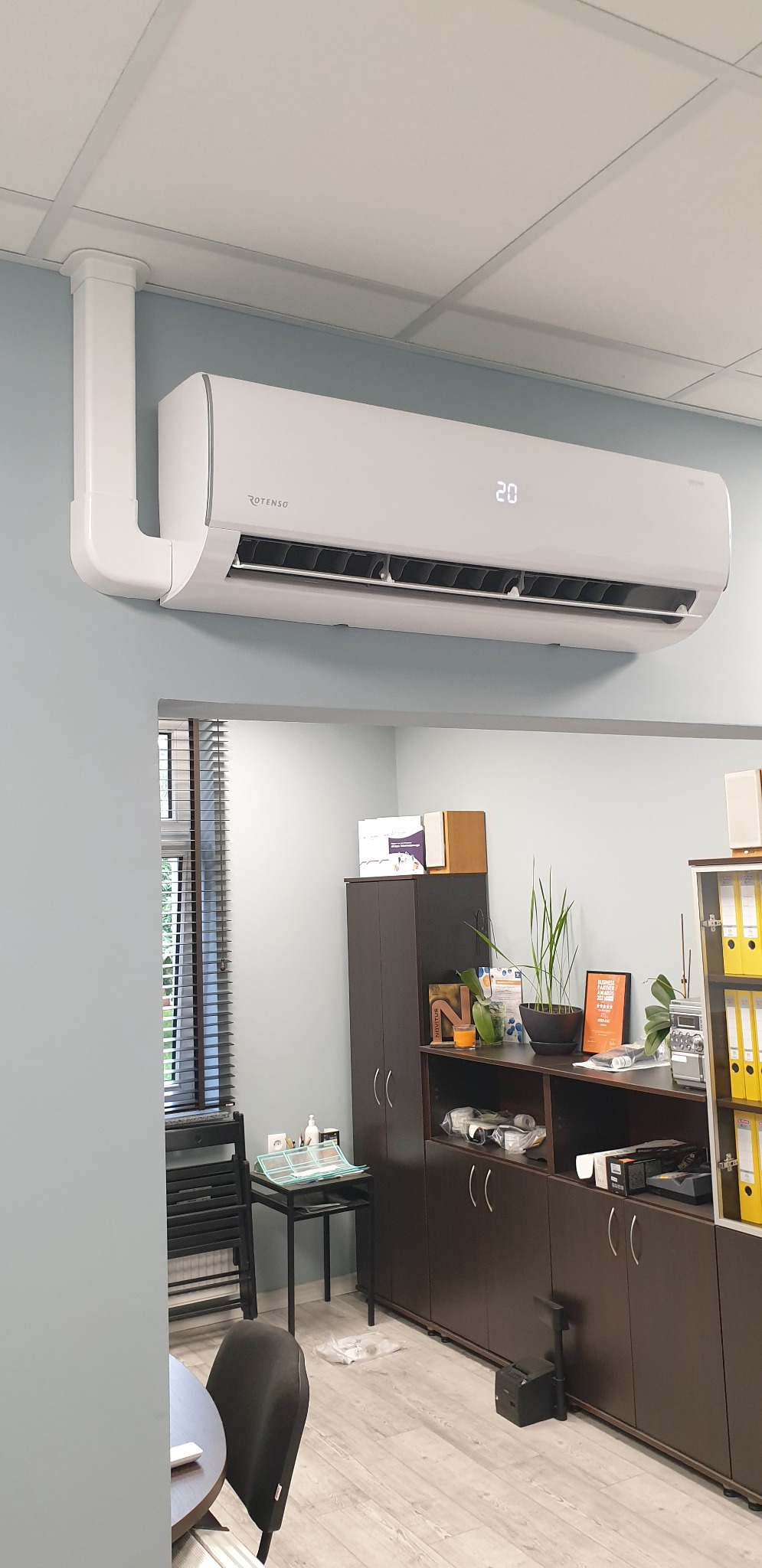 Biały klimatyzator Rotenso zamontowany na ścianie biura, widoczny wyświetlacz z temperaturą 20 stopni Celsjusza, białe rury instalacyjne prowadzone po suficie.
