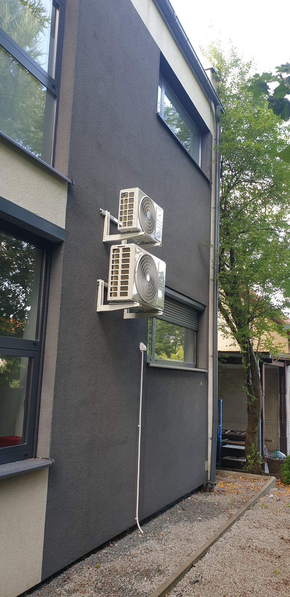 Dwa klimatyzatory zamontowane jeden nad drugim na elewacji domu pomalowanej na grafitowo, widoczne białe wsporniki i rurka odprowadzająca skropliny.