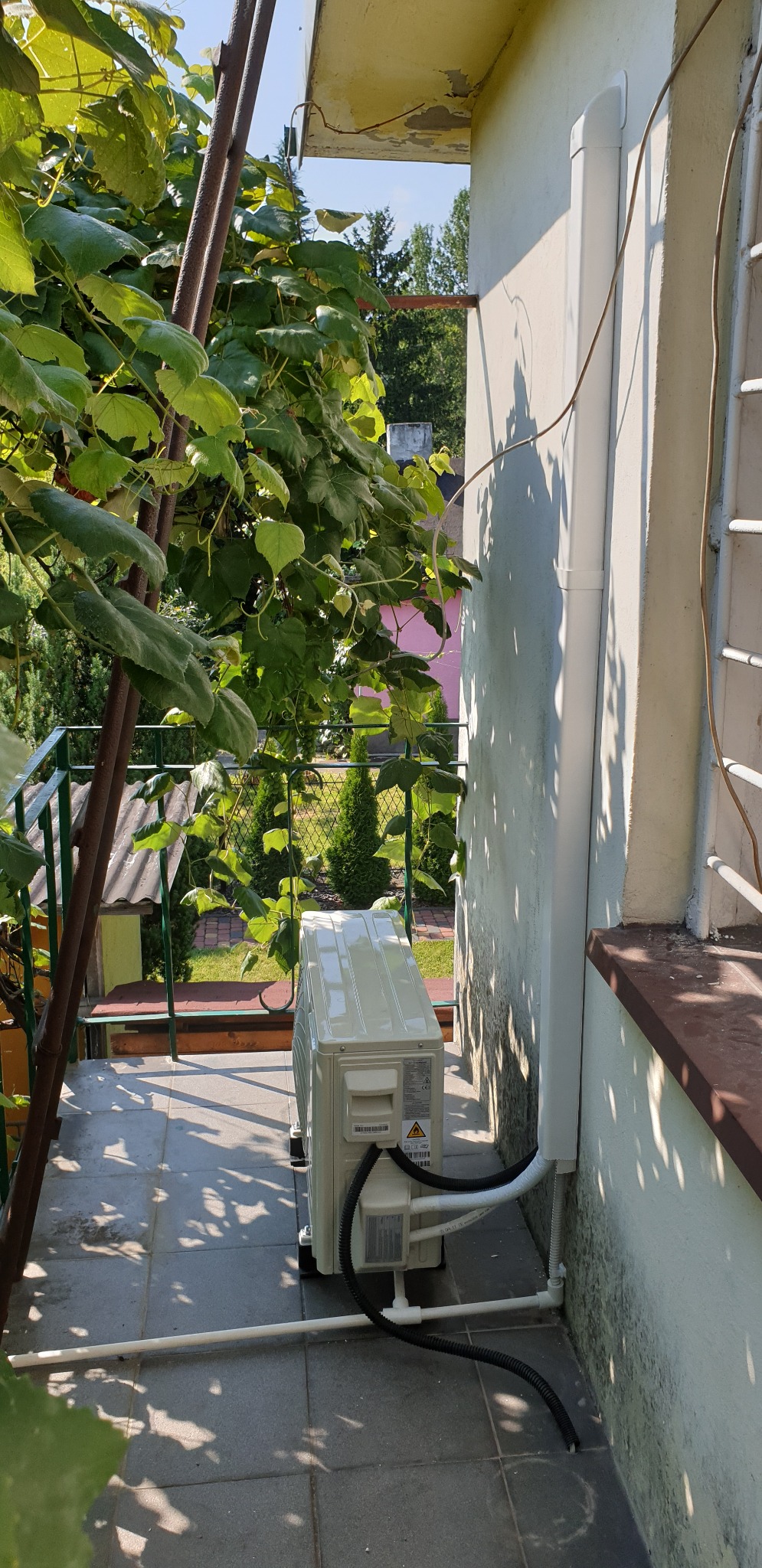 Zewnętrzna jednostka klimatyzacji zamontowana na balkonie domu, widoczne rury odprowadzające skropliny i przewody elektryczne, zieleń winorośli w tle.