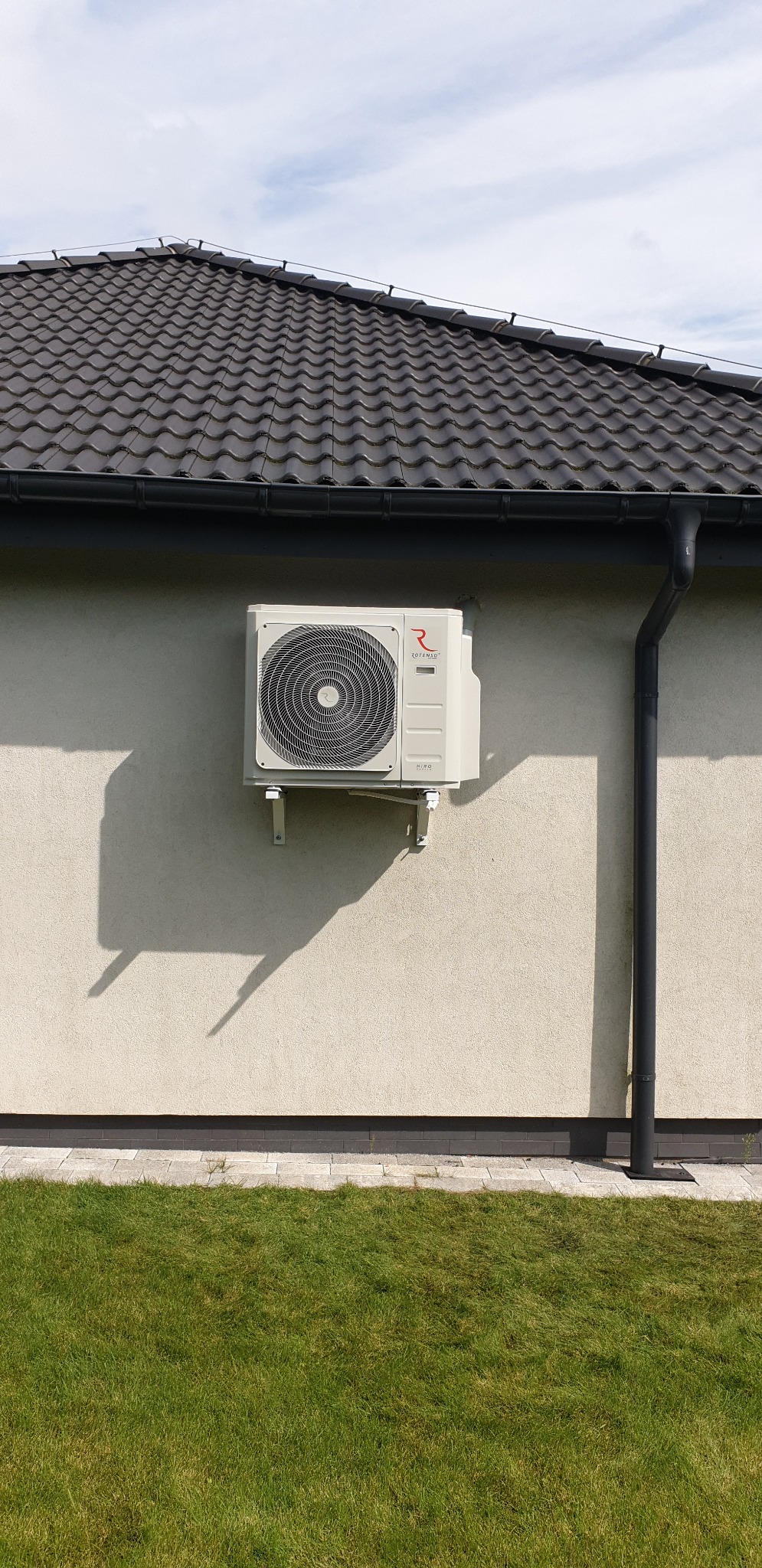 Zewnętrzna jednostka klimatyzacji marki Rotenso, zamontowana na elewacji jasnego domu jednorodzinnego z ciemnym dachem, widoczna z poziomu trawnika.