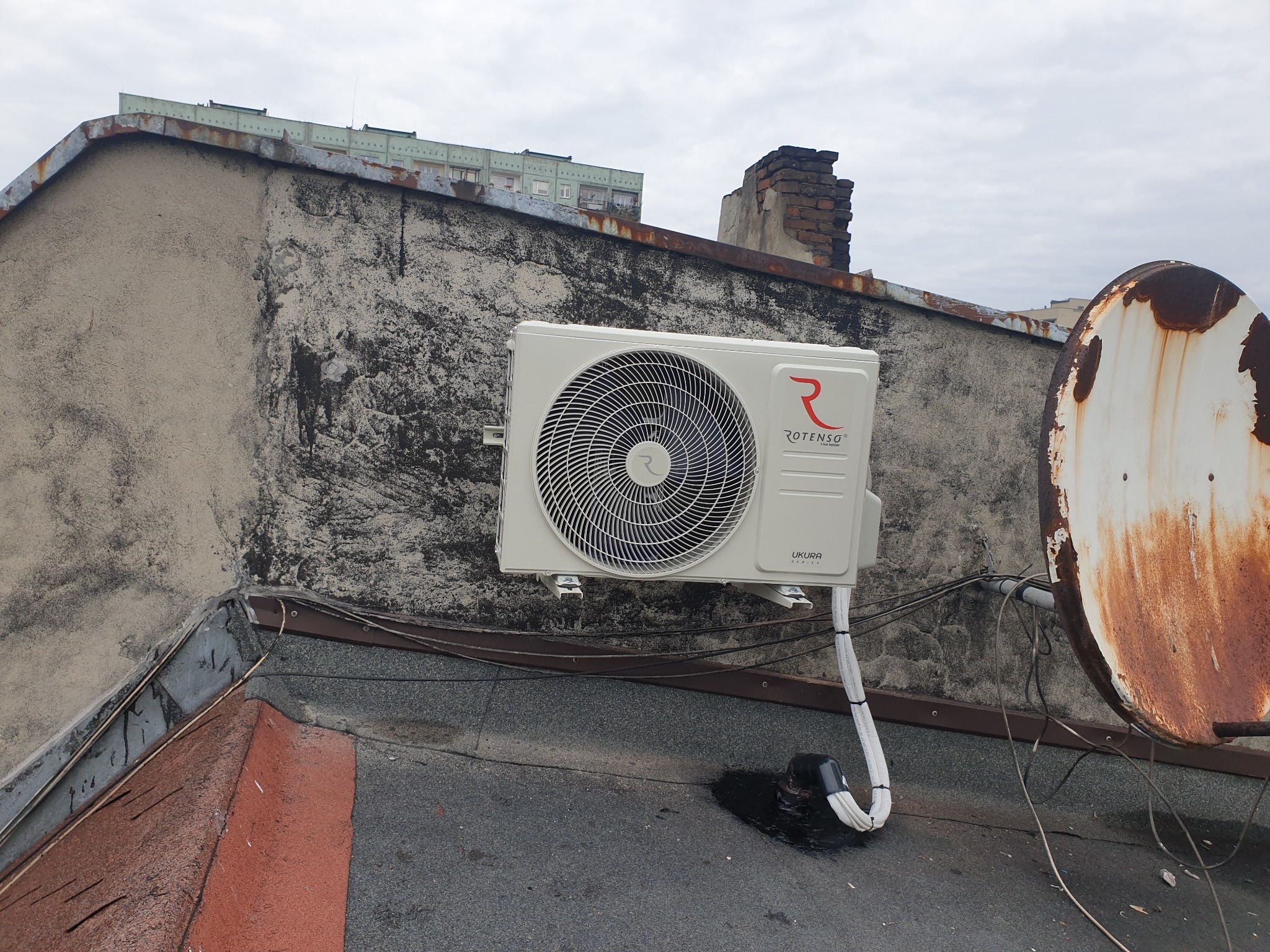 Biała jednostka zewnętrzna klimatyzacji Rotenso Ukura zamontowana na szczycie budynku z widoczną rdzewiejącą anteną satelitarną i zabrudzoną elewacją w tle.