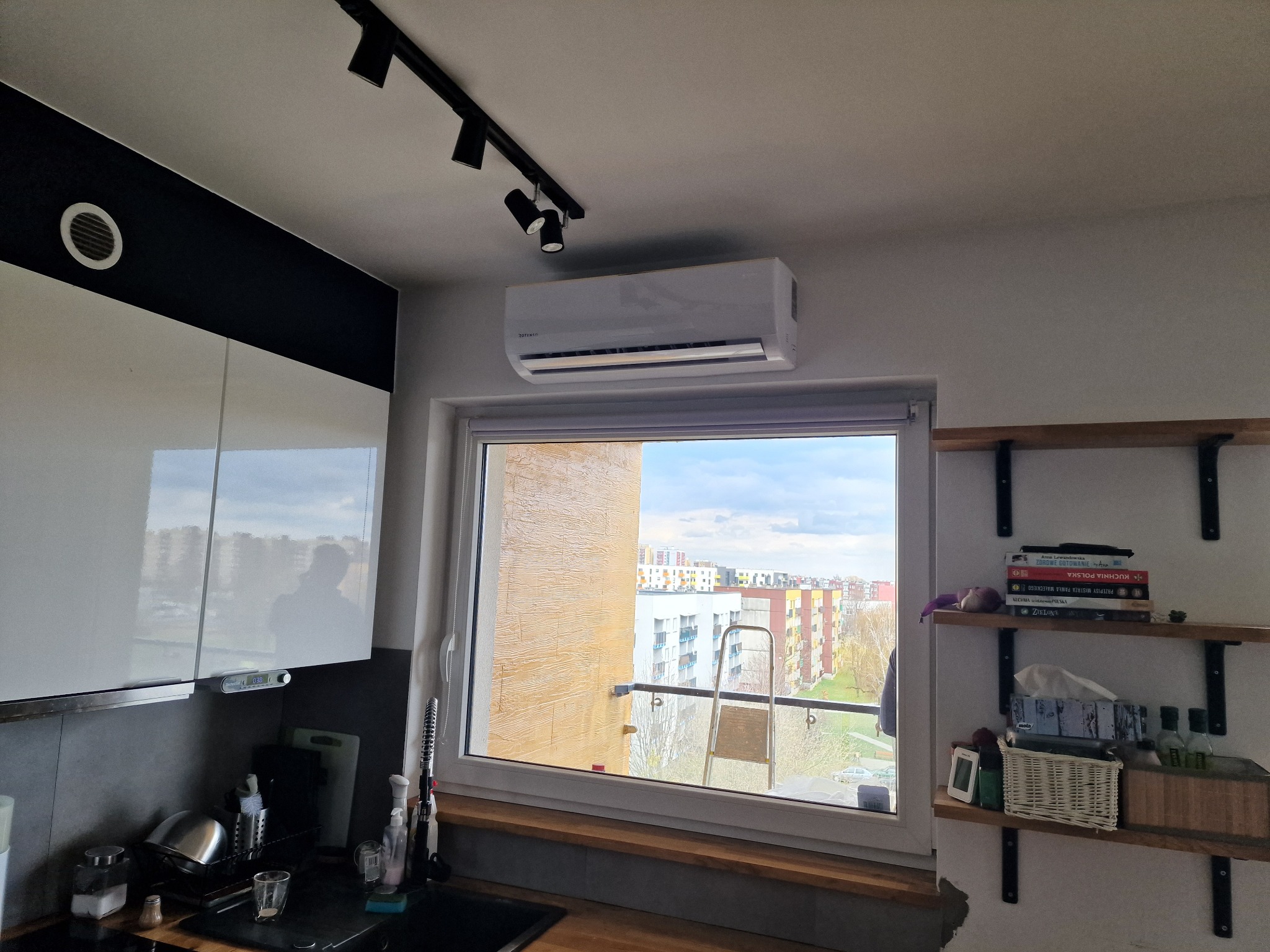 Biała jednostka klimatyzacyjna zamontowana nad oknem w mieszkaniu, widok na osiedle z drabinką na balkonie po drugiej stronie okna, oświetlenie punktowe na suficie.
