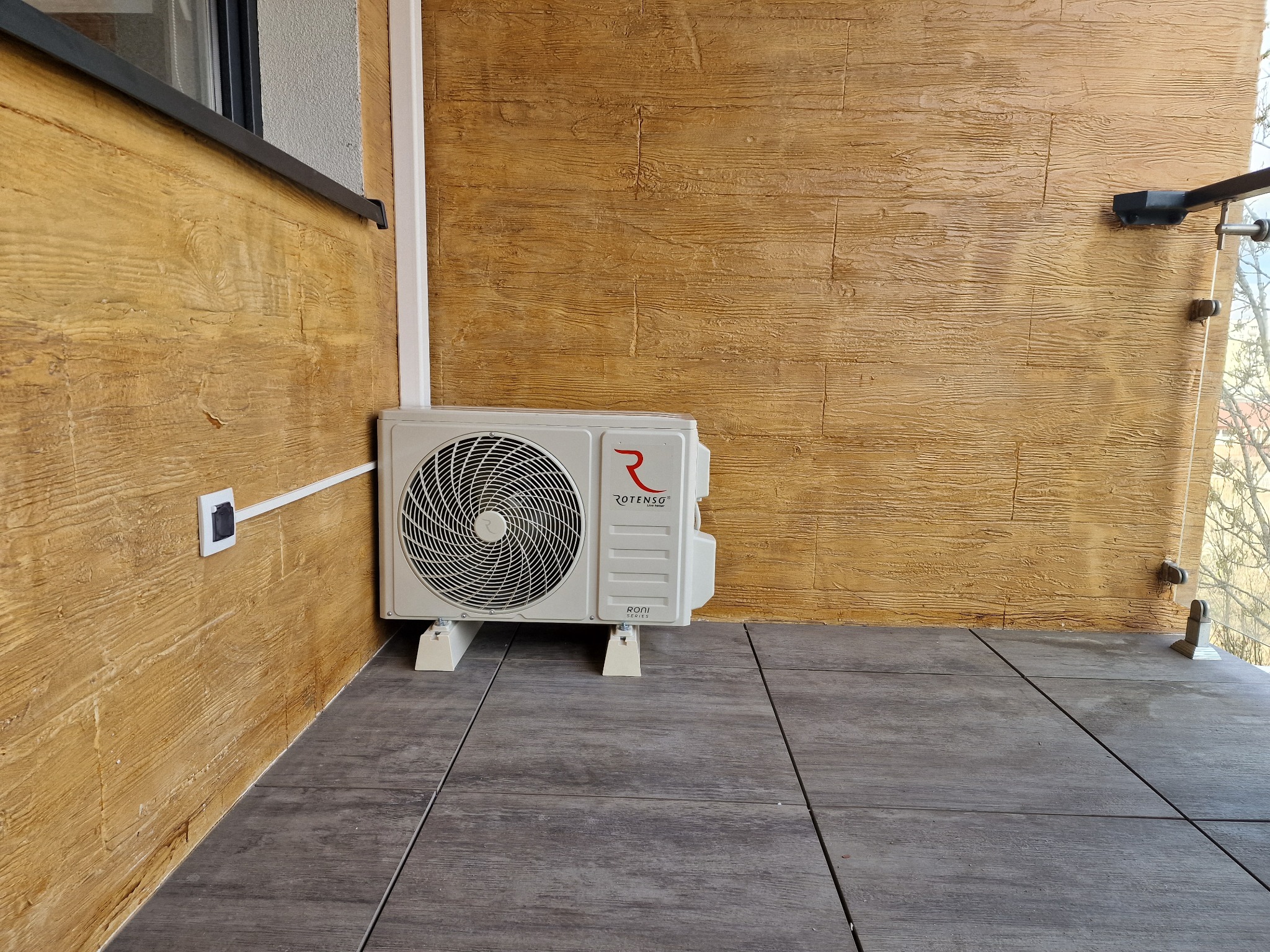 Zewnętrzna jednostka klimatyzacji Rotenso zamontowana na balkonie z drewnopodobną ścianą i szarą podłogą z płytek.