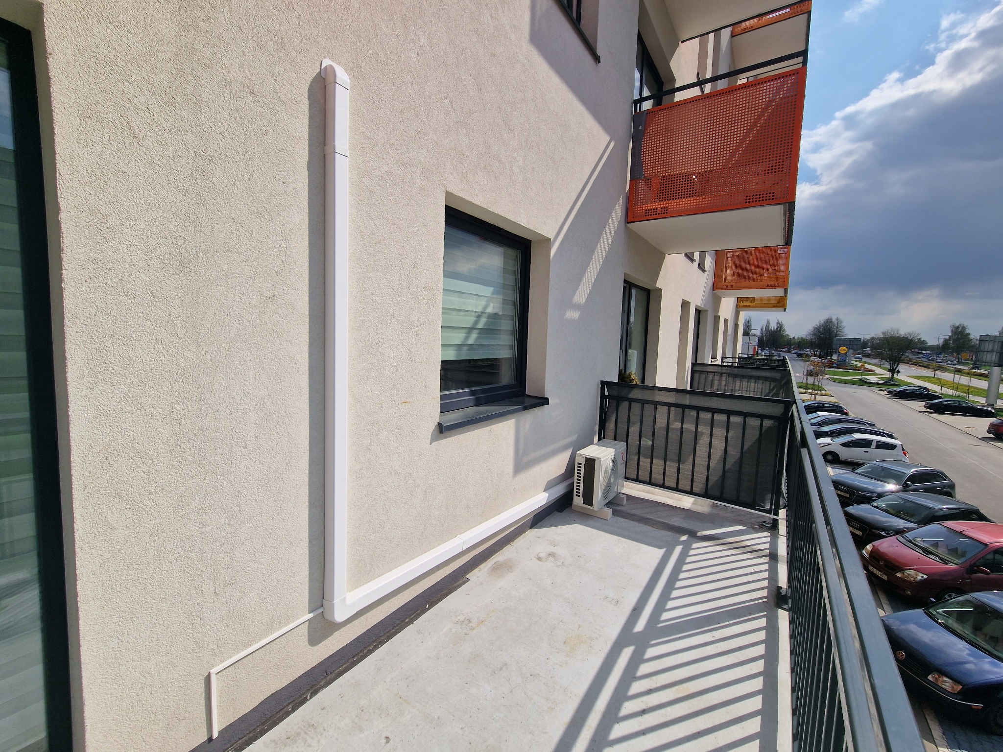 Kompaktowa jednostka zewnętrzna klimatyzacji zamontowana na balkonie nowoczesnego apartamentowca, z widocznymi białymi maskownicami przewodów na elewacji.