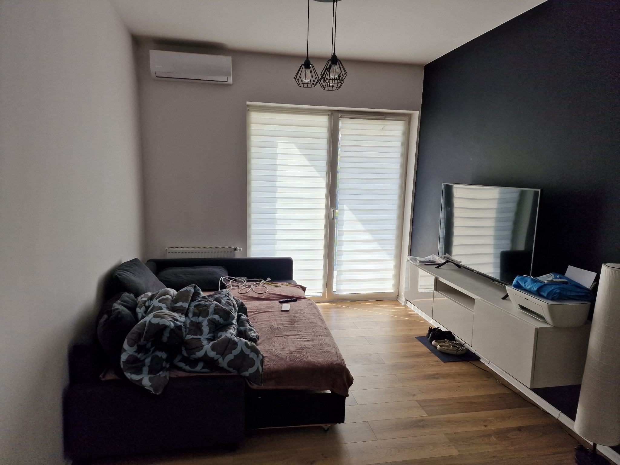 Wnętrze pokoju dziennego z klimatyzatorem na ścianie, widok na sofę, okno z żaluzjami, telewizor i białą szafkę RTV.