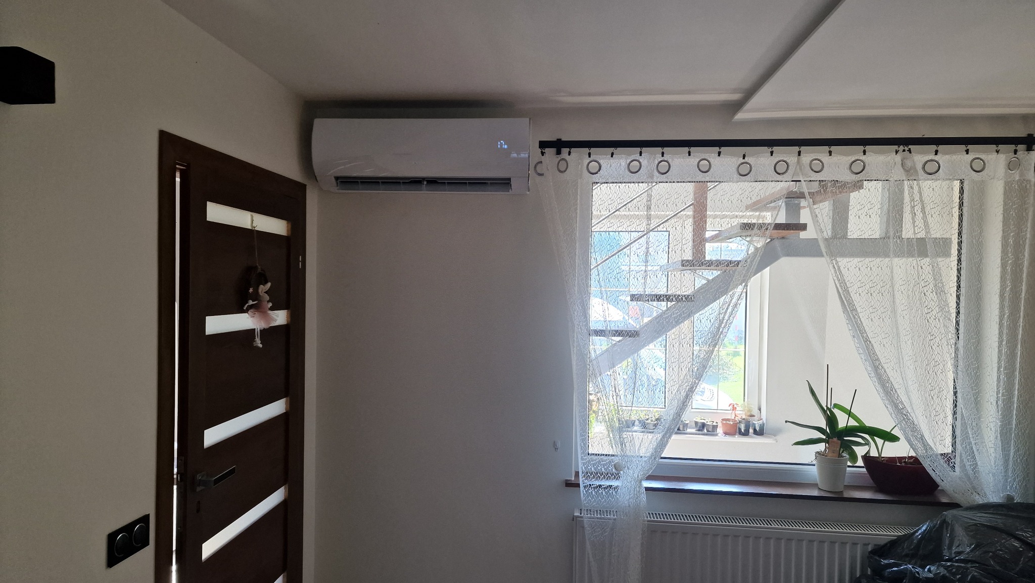 Biały klimatyzator ścienny zamontowany w jasnym pokoju, widok na okno z firanką i schodami w tle.