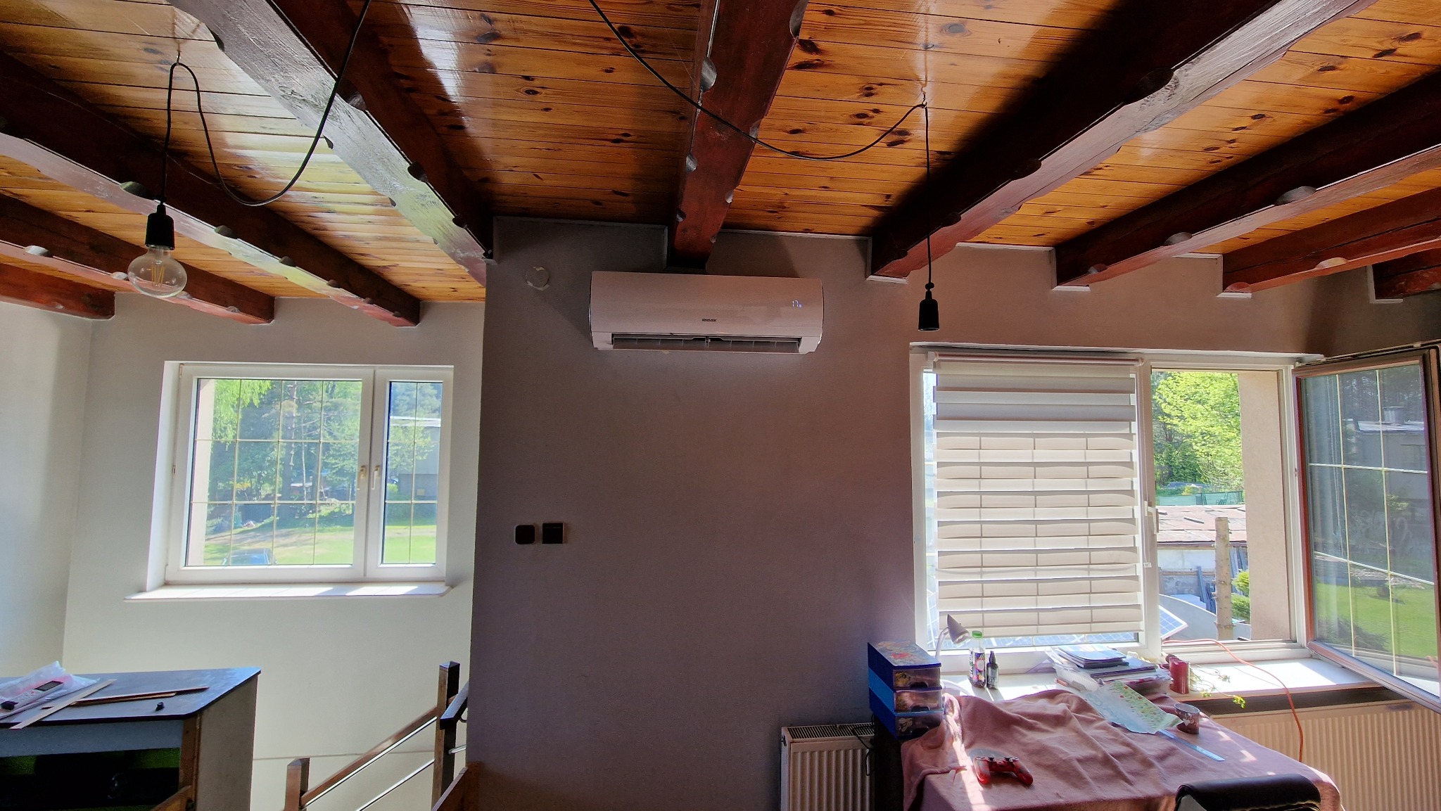 Wnętrze pokoju z zamontowanym białym klimatyzatorem na szarej ścianie, widoczne drewniane belki stropowe, okno z widokiem na zieleń, oraz wisząca żarówka na kablu.