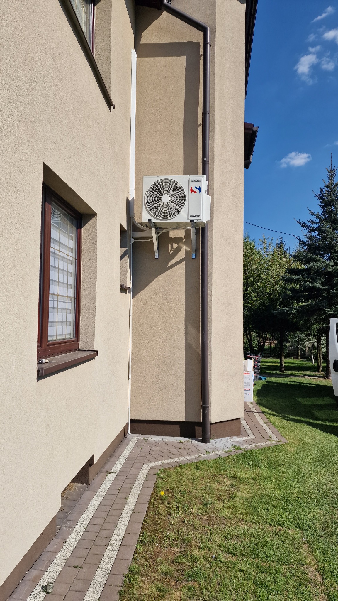 Zewnętrzna jednostka klimatyzacji Sinclair zamontowana na elewacji domu z brązową rynną i oknem w tle, na zewnątrz kostka brukowa i trawnik.