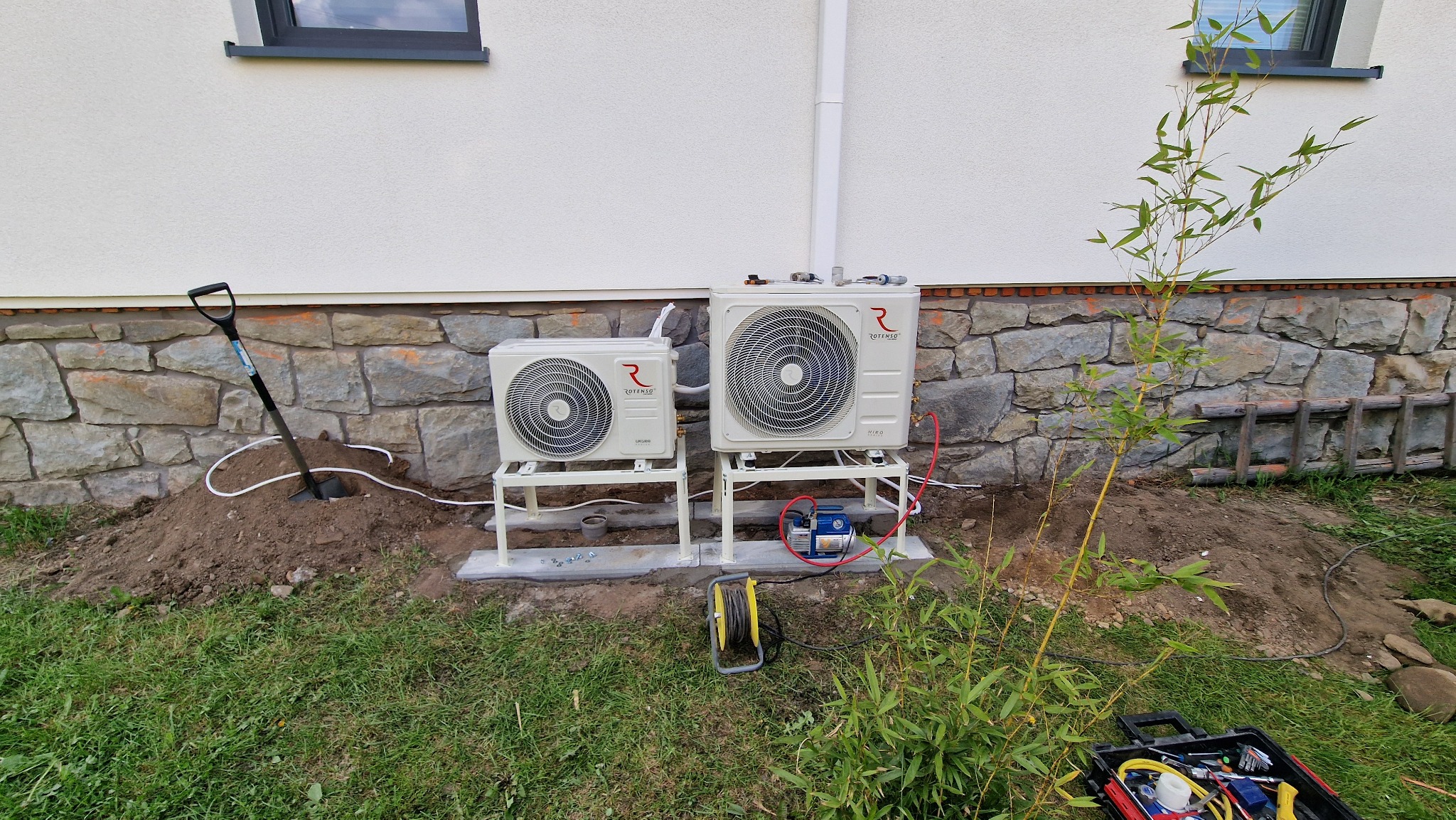 Dwa klimatyzatory typu split, marki Rotenso, zamontowane na metalowych stelażach przed budynkiem z kamienną podmurówką; widoczna koparka, narzędzia i sprzęt do serwisu klimatyzacji.