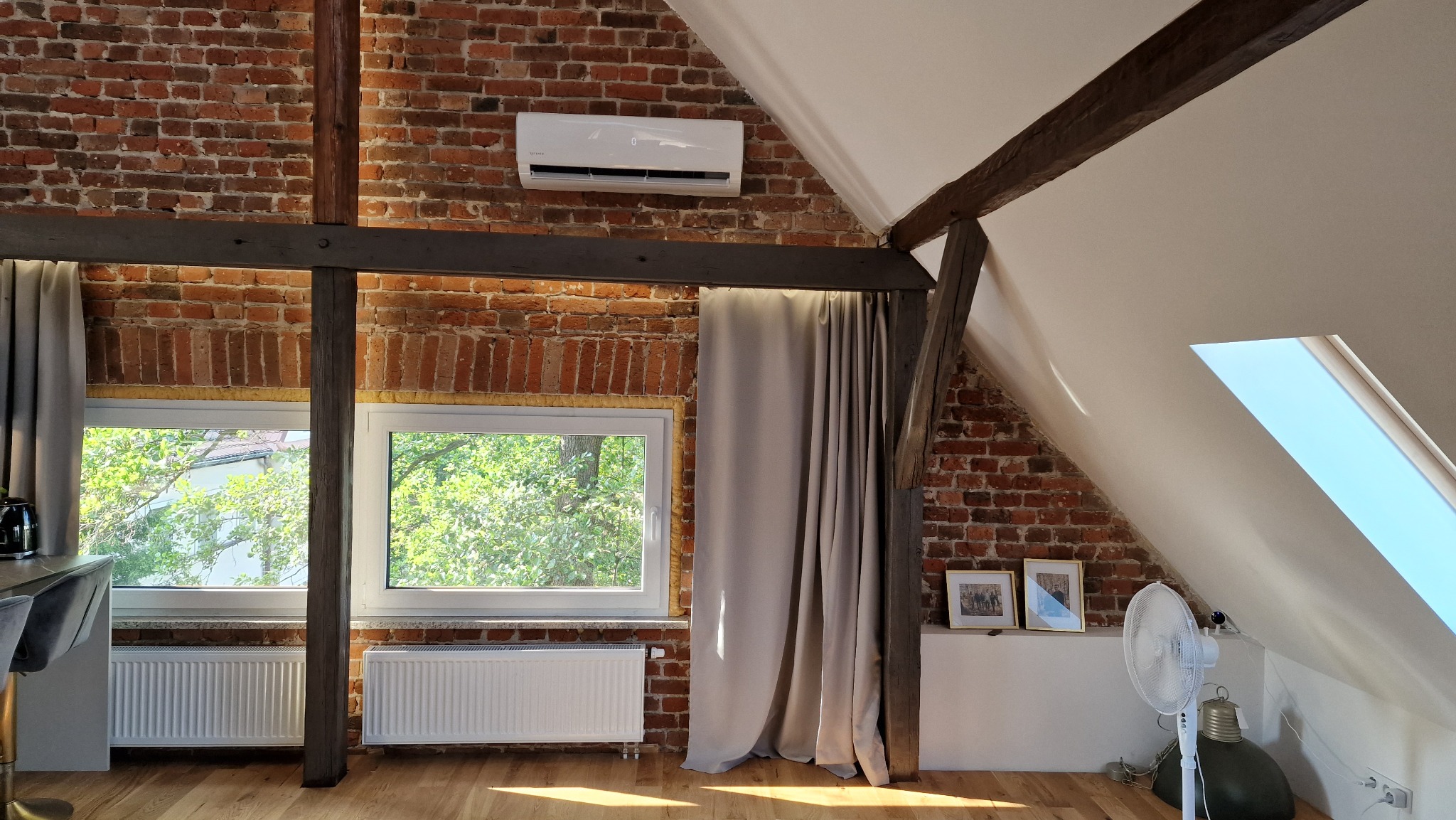 Klimatyzator ścienny zamontowany na ceglanej ścianie poddasza z drewnianymi belkami stropowymi i oknami.