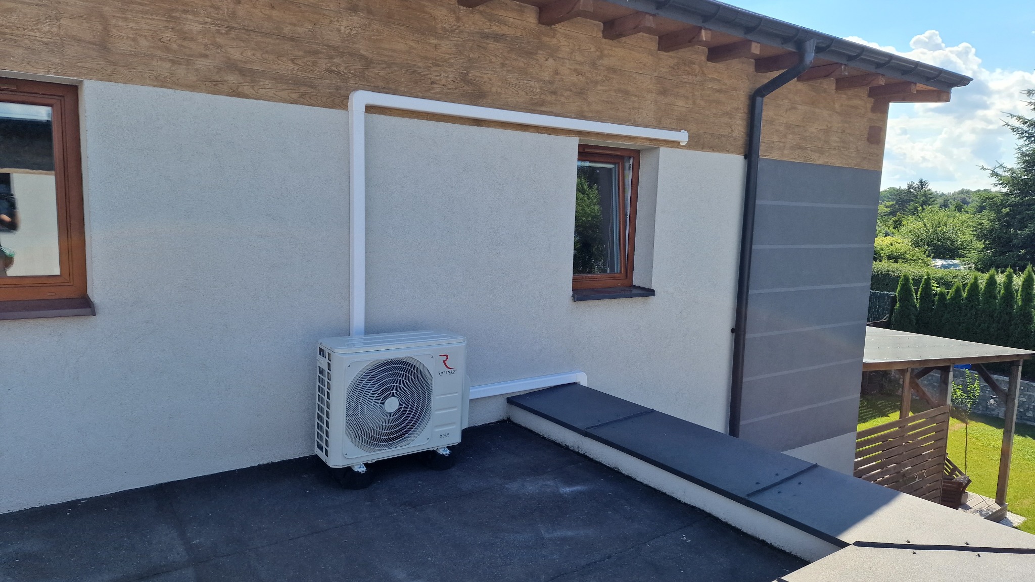 Zewnętrzna jednostka klimatyzacji zamontowana na dachu domu, z widocznymi rurami odprowadzającymi i elementami mocującymi.